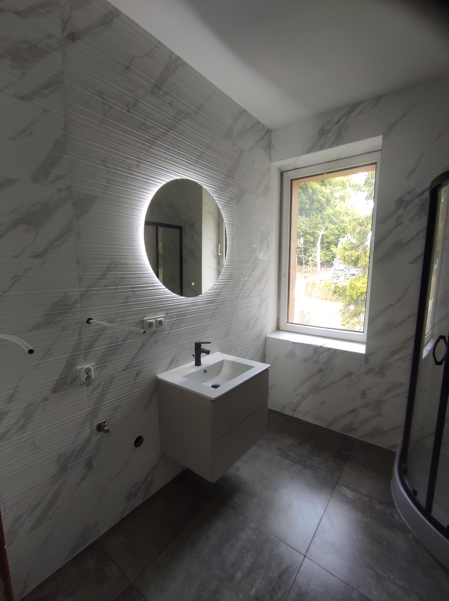 Nowoczesna łazienka z biało-szarymi płytkami imitującymi marmur, okrągłe lustro z podświetleniem LED, wisząca szafka z umywalką i fragment kabiny prysznicowej.