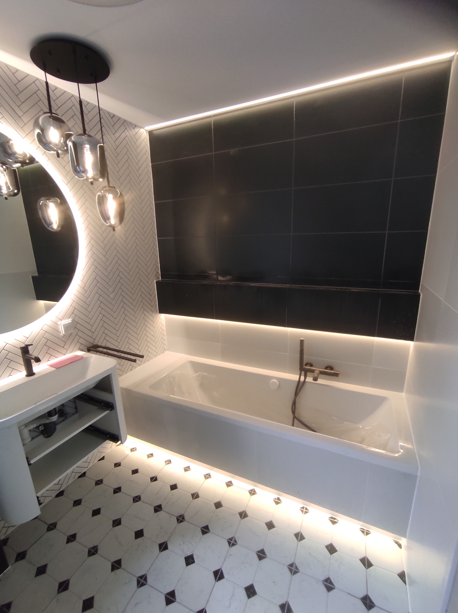 Nowoczesna łazienka z czarno-białymi płytkami, podświetlaną wanną i okrągłym lustrem z lampami wiszącymi.