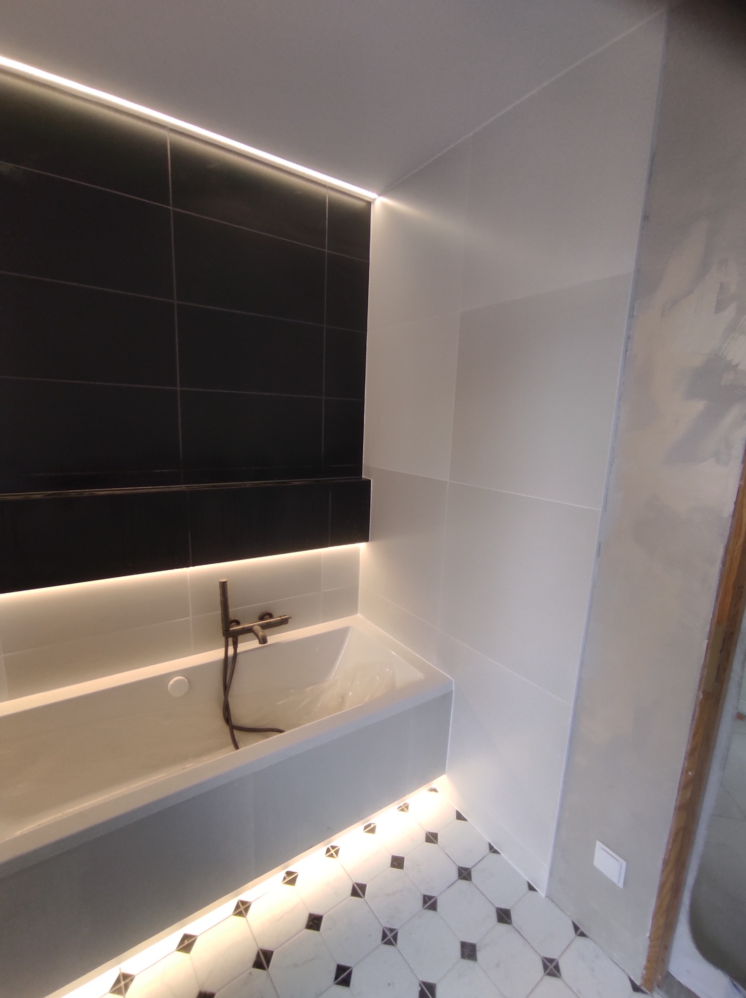 Nowoczesna łazienka z czarno-białymi płytkami podłogowymi, podświetlaną wanną i czarnymi płytkami na ścianie z listwą LED, bateria w stylu retro.