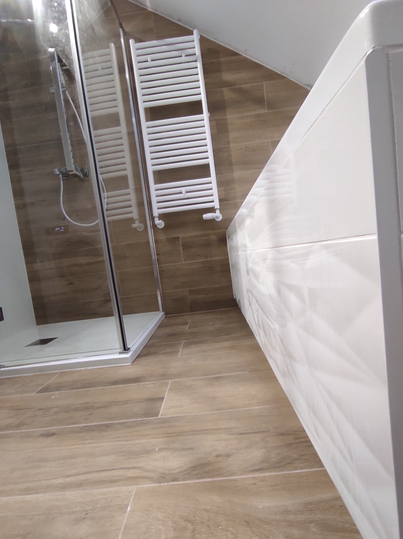 Nowoczesna łazienka z kabiną prysznicową typu walk-in, grzejnikiem drabinkowym i umywalką z szafką w kolorze białym, wykończona płytkami imitującymi drewno.