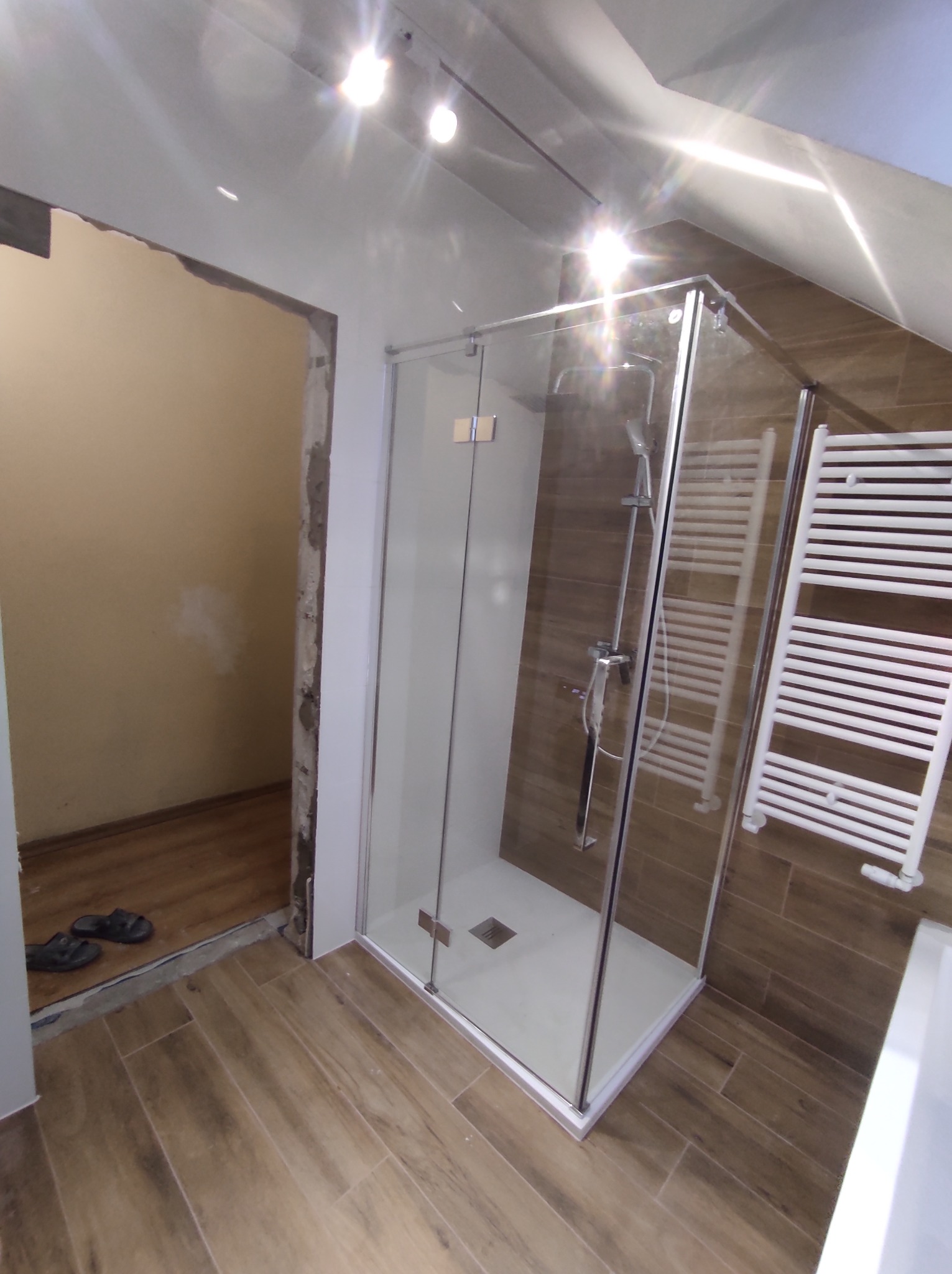 Montaż kabiny prysznicowej w łazience z brązowymi płytkami imitującymi drewno, widoczne niedokończone wykończenie w przejściu do innego pomieszczenia i biały grzejnik drabinkowy.