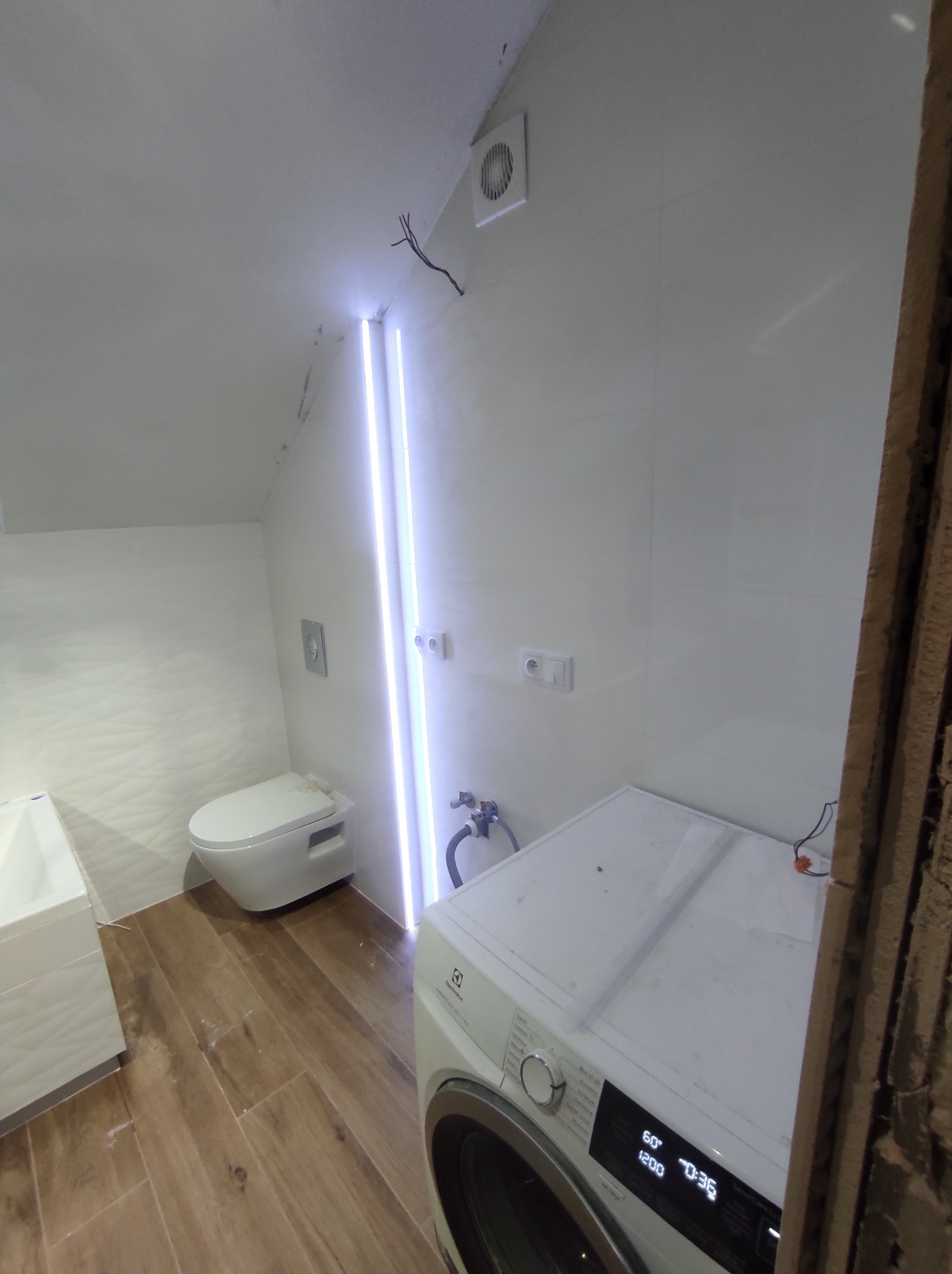 Ukończona łazienka na poddaszu z drewnopodobną podłogą, białą armaturą i pralką Electrolux. Ściany wyłożone białymi płytkami z subtelnym wzorem, oświetlenie LED w pionowej szczelinie. Widoczne...