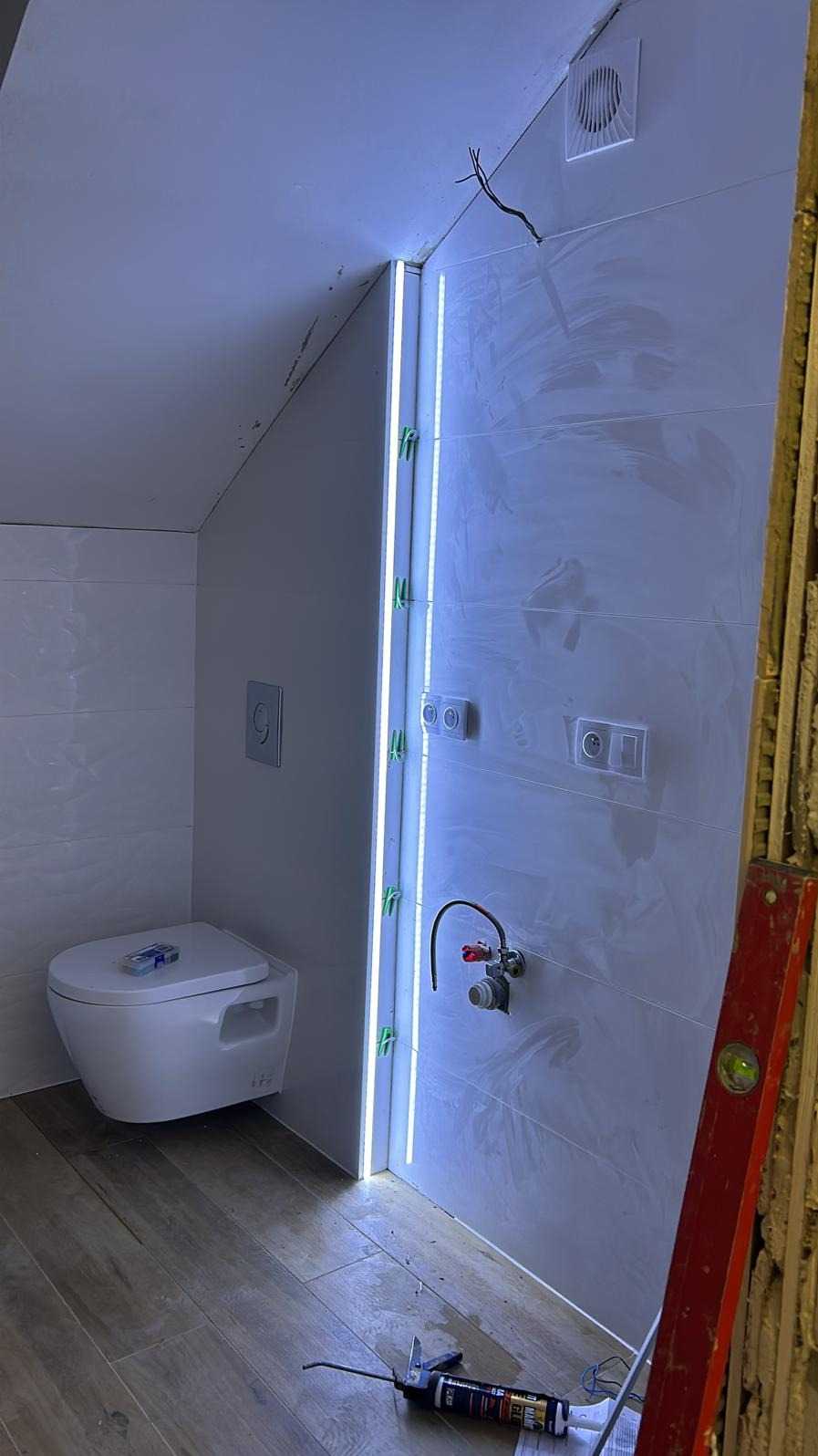 Świeżo wykafelkowana łazienka na poddaszu z podwieszaną toaletą, listwą LED i niedokończonymi detalami, takimi jak wystające kable i poziomica oparta o ścianę.