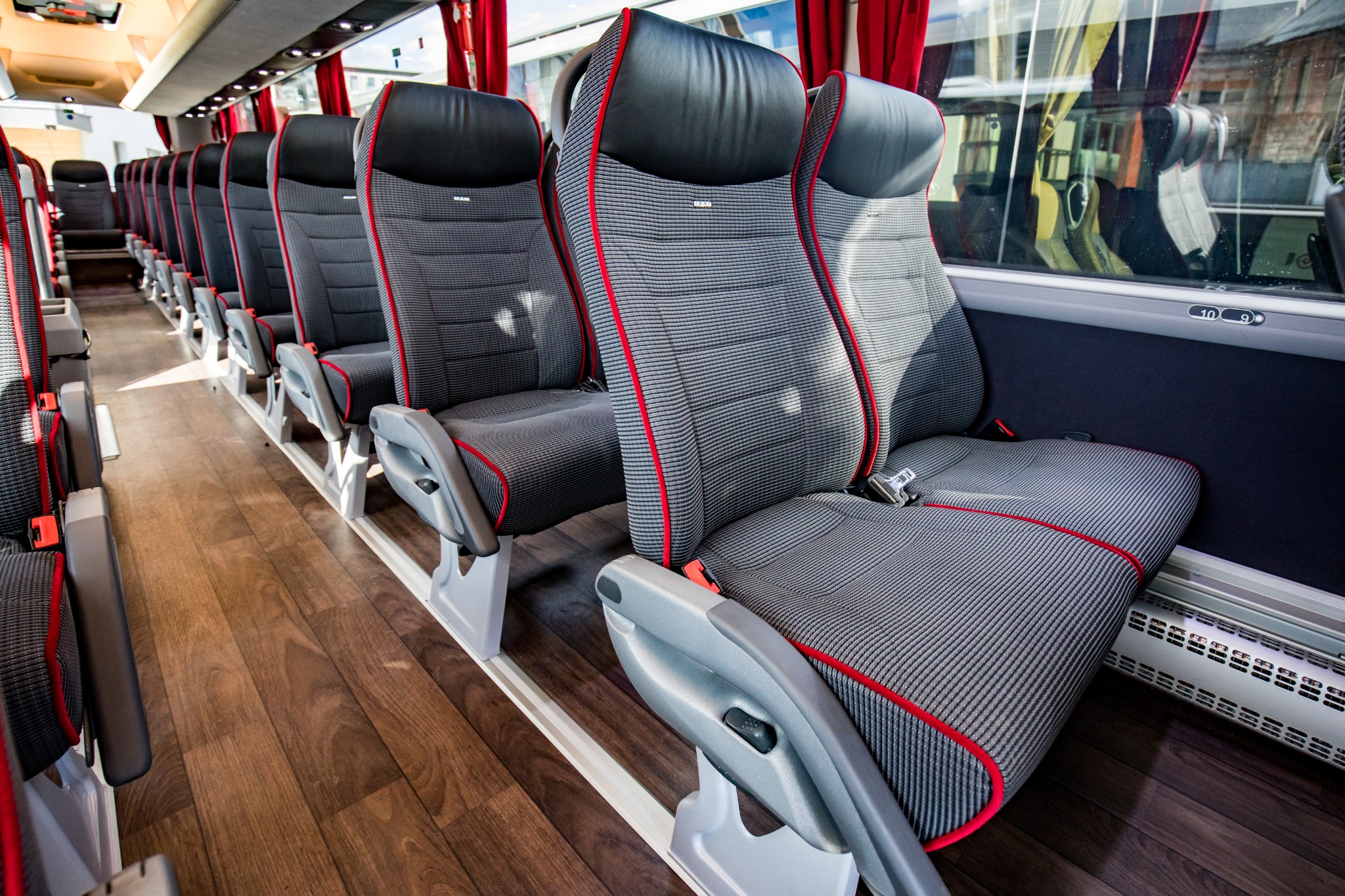 Wnętrze autobusu turystycznego: rzędy szarych foteli z czerwonymi lamówkami, brązowa podłoga imitująca drewno, widoczne zasłonki w oknach i numery miejsc nad oknem.