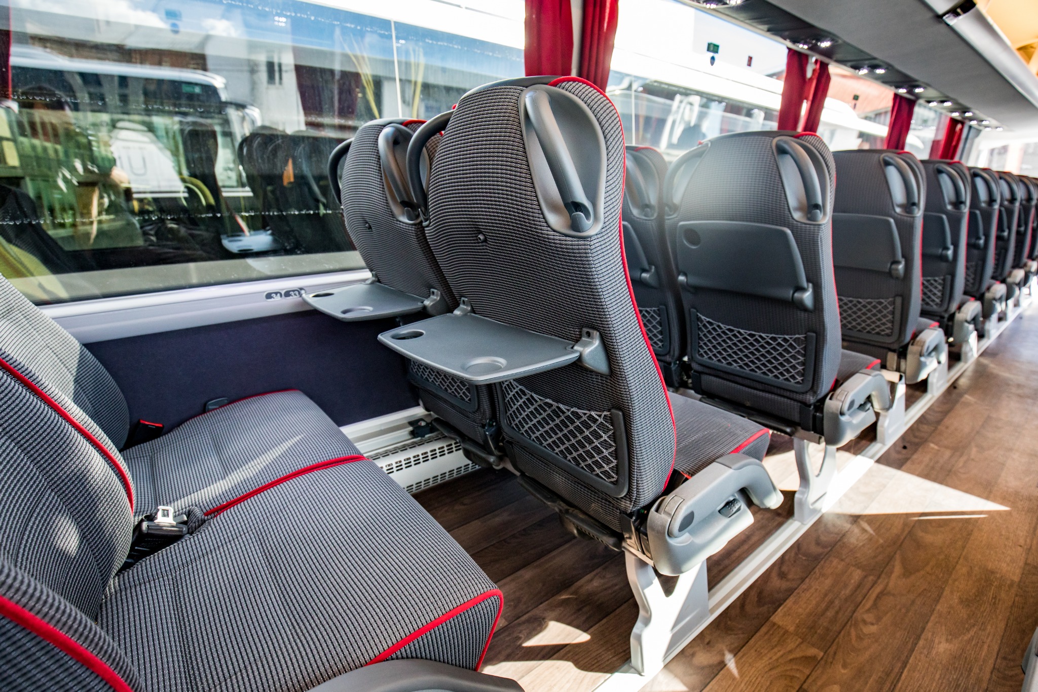Wnętrze autobusu turystycznego z rzędem szarych foteli z czerwonymi lamówkami, rozkładanymi stolikami i siatkami na bagaż, widok z perspektywy pasażera.