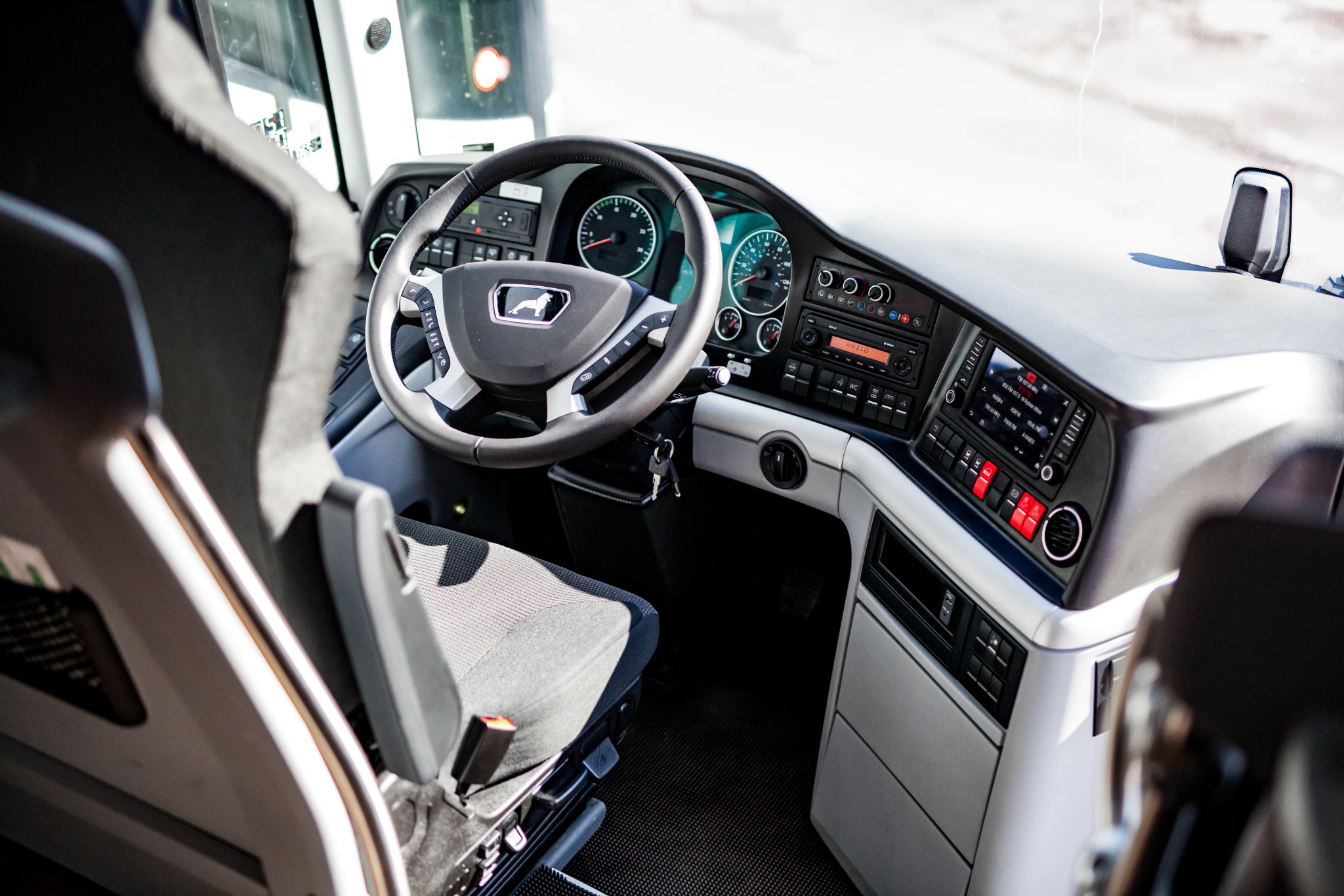 Wnętrze kabiny autobusu MAN z widocznym kokpitem, kierownicą z logo lwa i fotelem kierowcy z pomarańczowym pasem bezpieczeństwa.