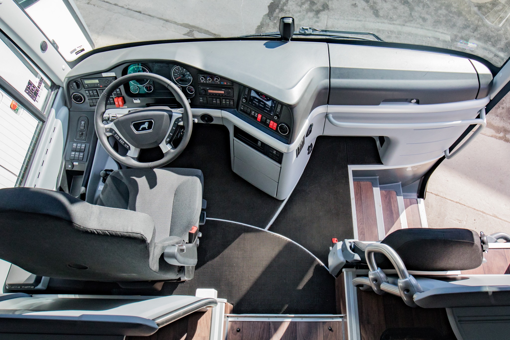 Wnętrze autobusu z widokiem na kokpit kierowcy, szare fotele, deska rozdzielcza z logo MAN i schodki wejściowe.