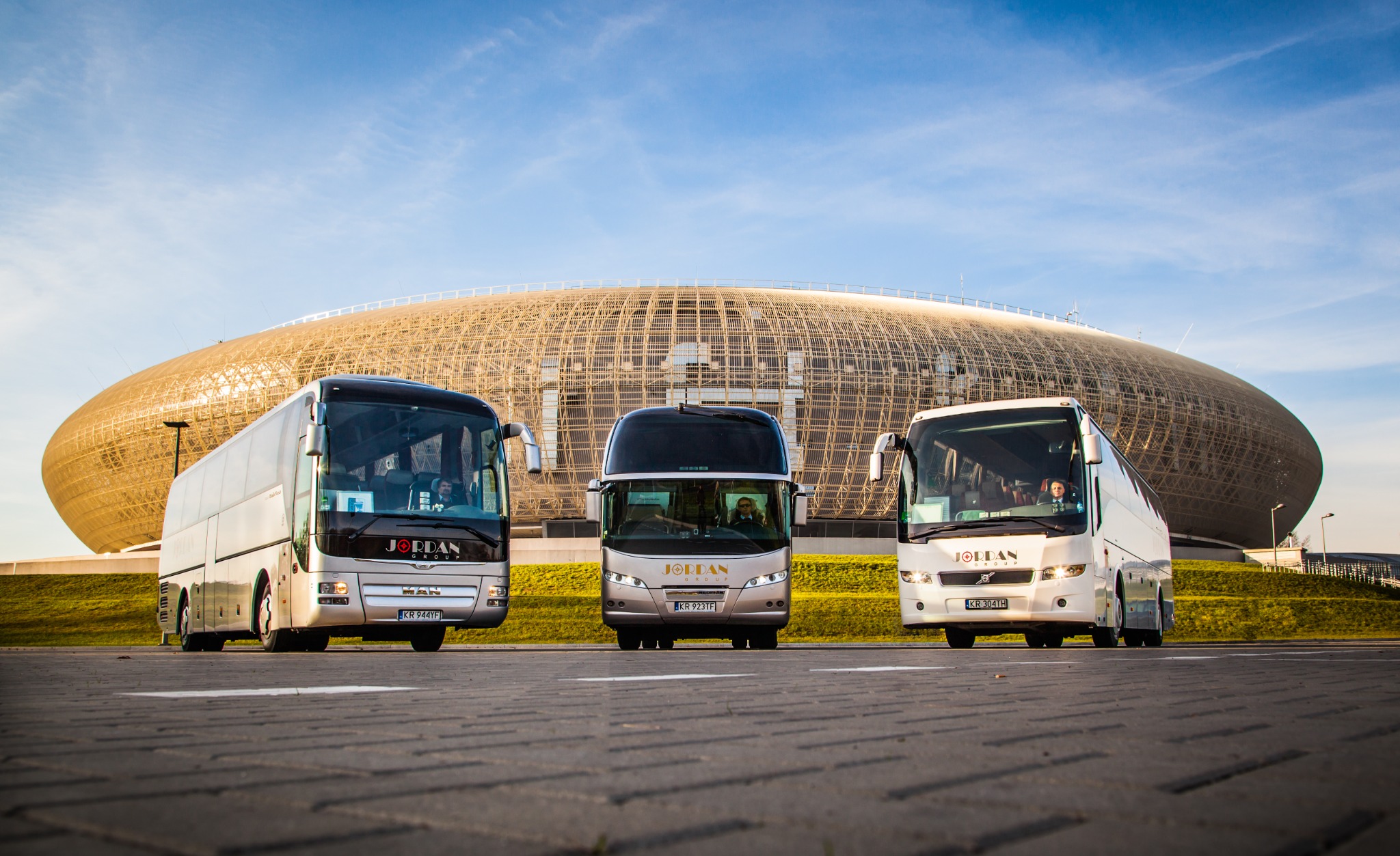 Trzy autokary firmy transportowej Jordan Group ustawione na parkingu przed Stadionem Energa Gdańsk w słoneczny dzień.