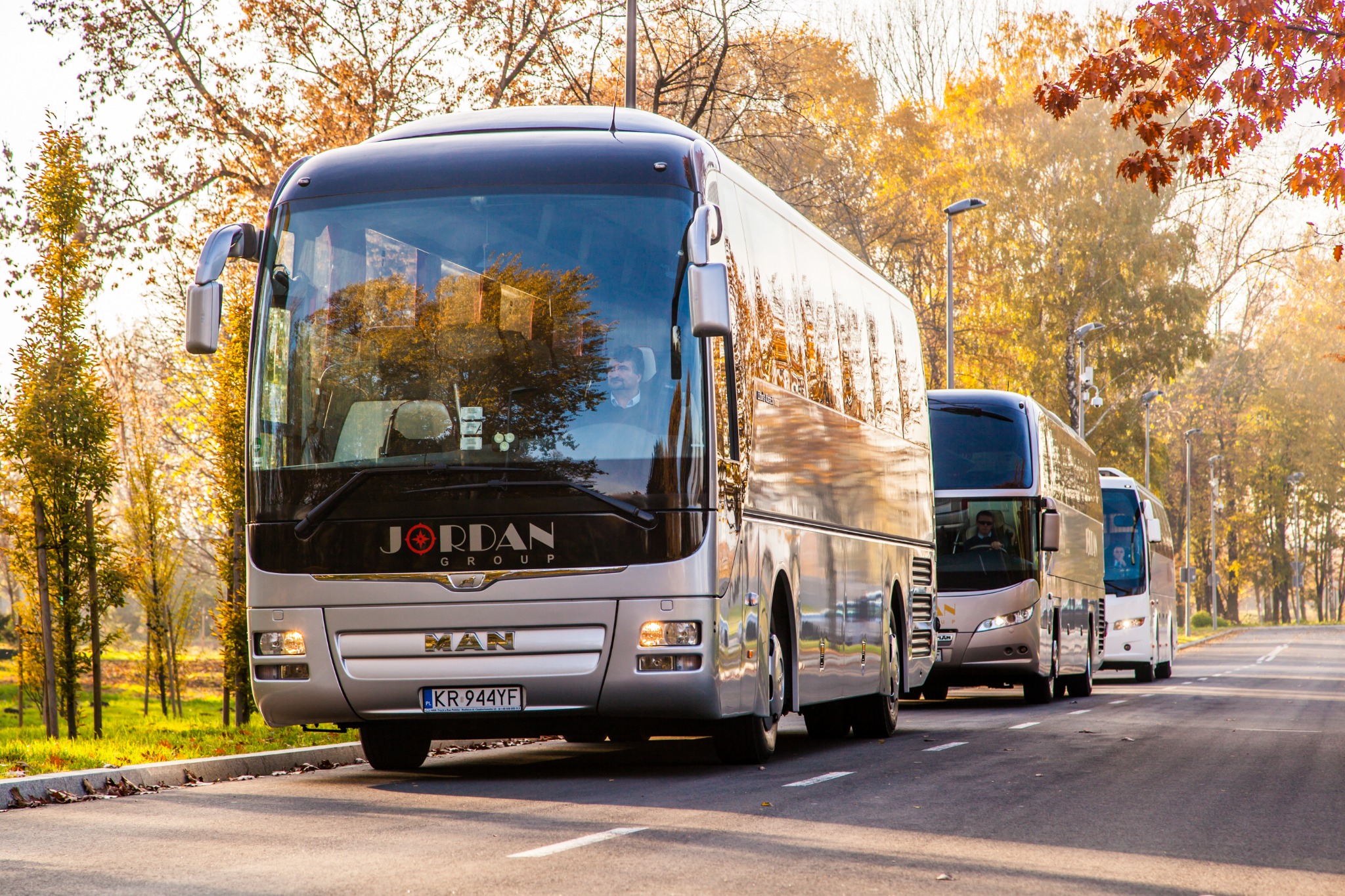 Srebrny autokar MAN Jordan Group w jesiennej scenerii, z widocznym kierowcą i tablicą rejestracyjną KR 944YF, ustawiony w rzędzie z innymi autokarami na drodze w parku.