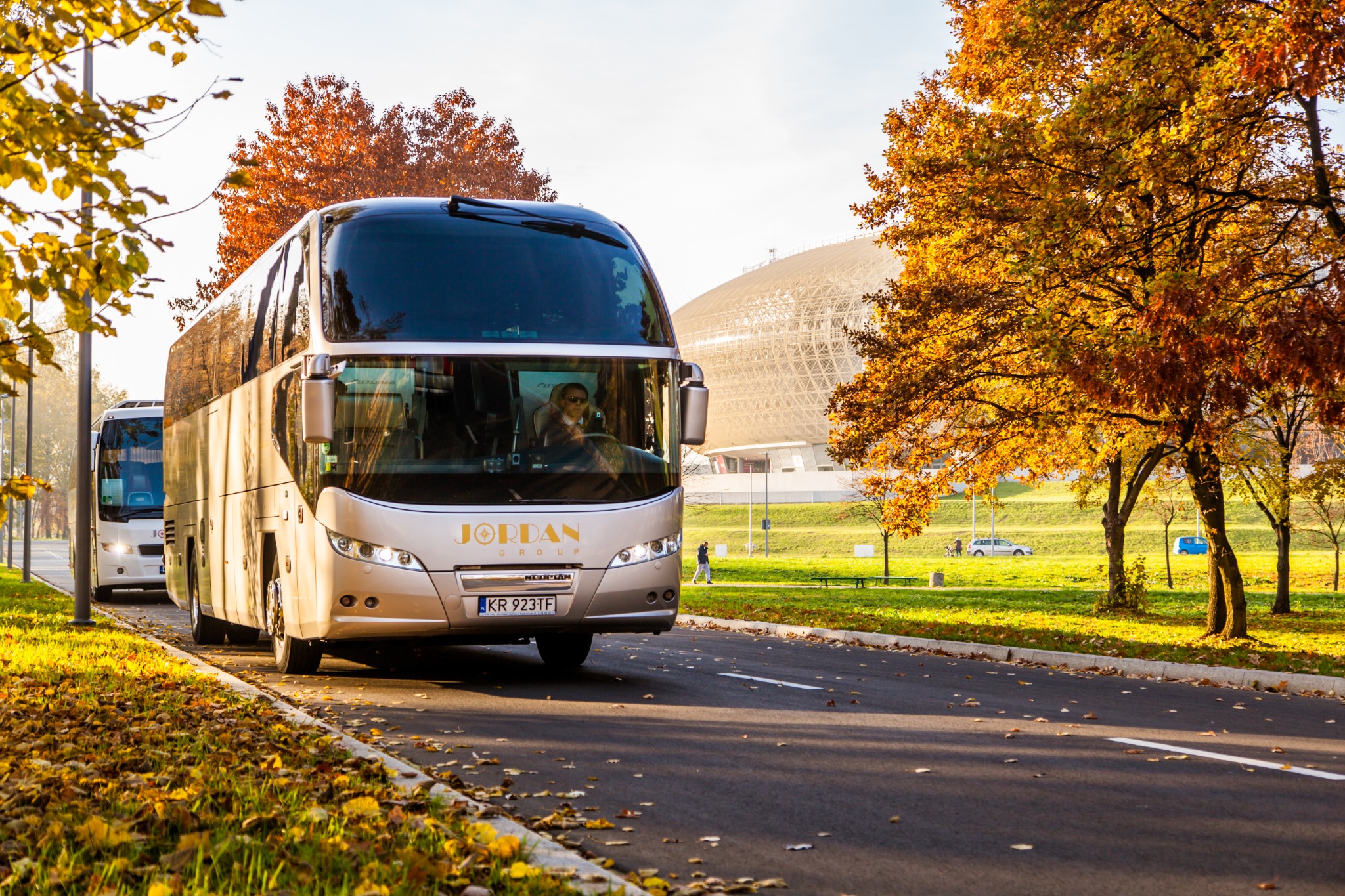 Beżowy autokar Jordan Group na drodze, w tle hala widowiskowa w Krakowie, jesienne drzewa