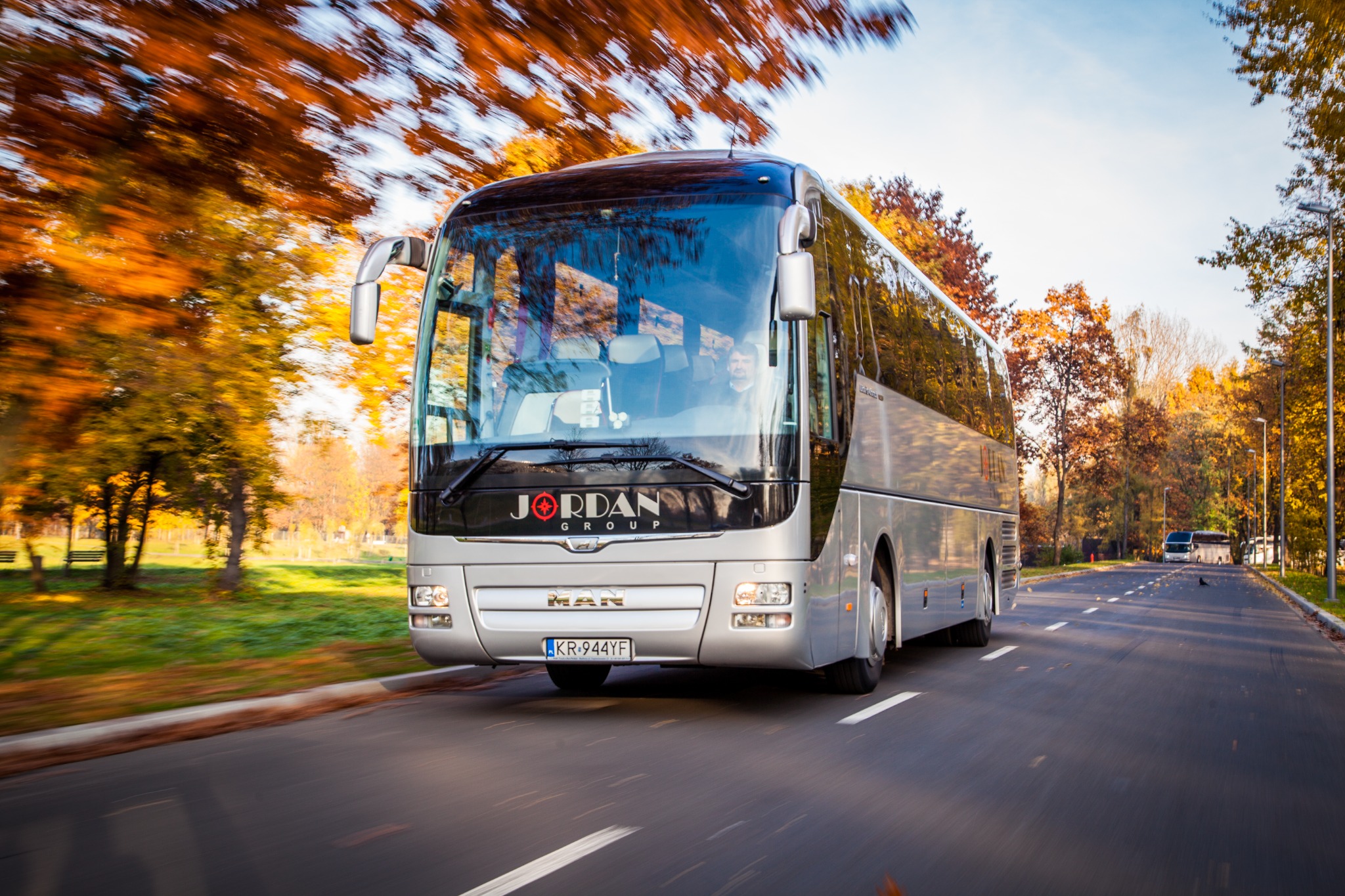 Srebrny autokar Jordan Group MAN w ruchu na asfalcie, jesienne drzewa w tle, kierowca widoczny przez szybę.
