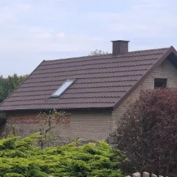 Dom z brązowym dachem z dachówki trapezowej i oknem dachowym, komin z obróbką blacharską. Budynek otoczony zielenią w słoneczny dzień.