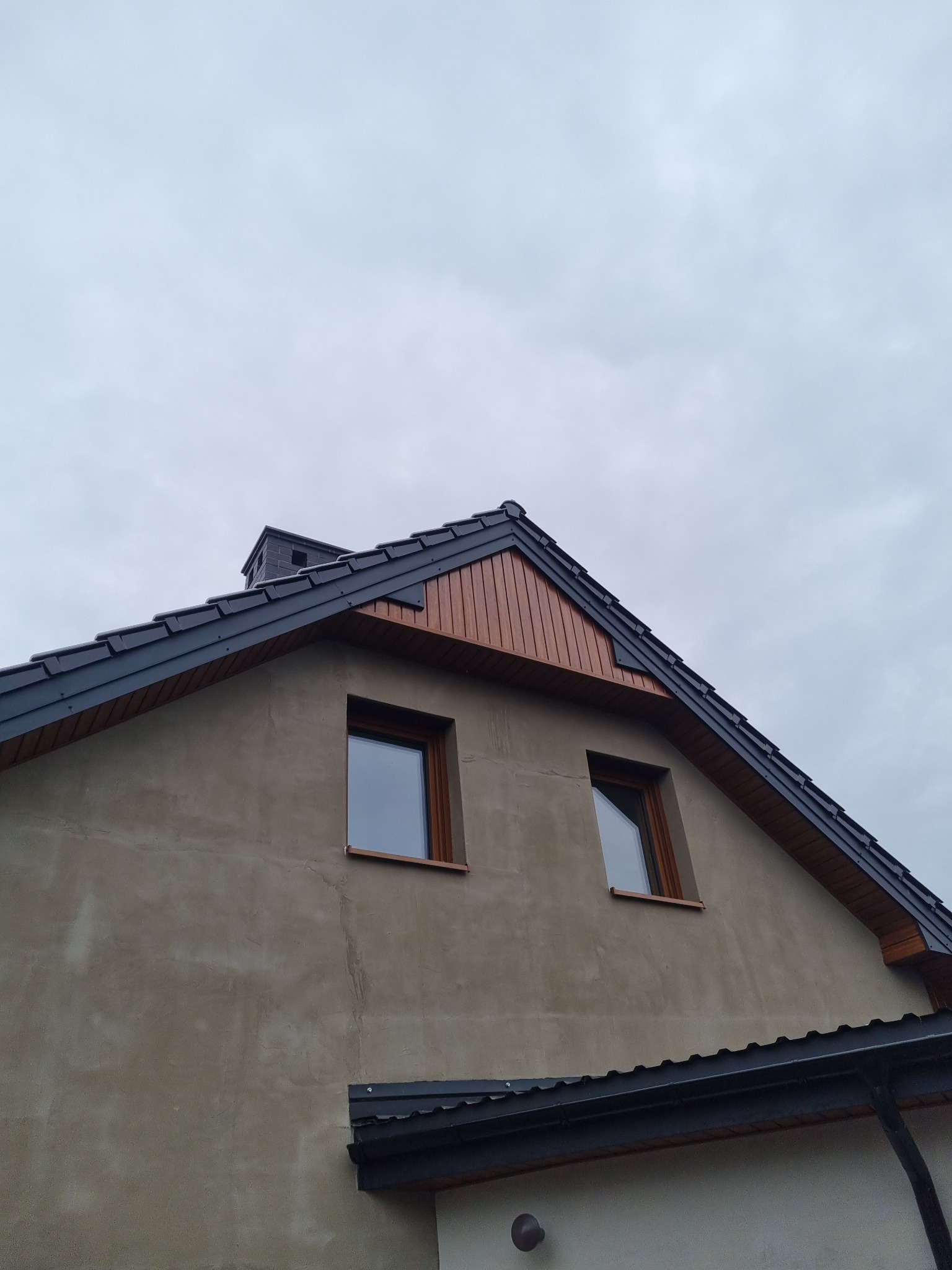 Fragment domu z ciemnoszarym dachem z dachówki ceramicznej, drewnianą podbitką i dwoma oknami na poddaszu, sfotografowany pod pochmurnym niebem.