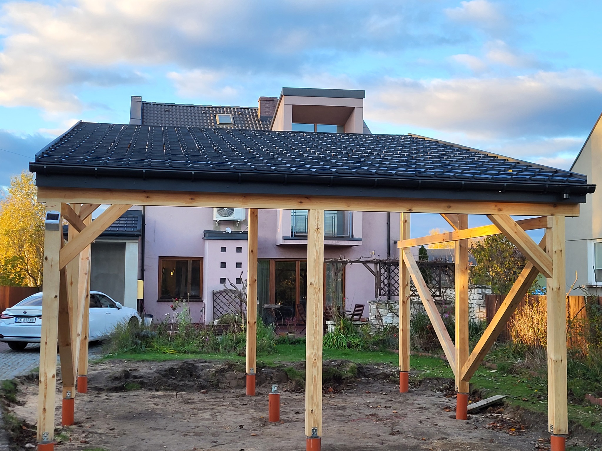 Drewniana konstrukcja z czarnym dachem z dachówki ceramicznej w trakcie budowy, widok na dom i ogród w tle.