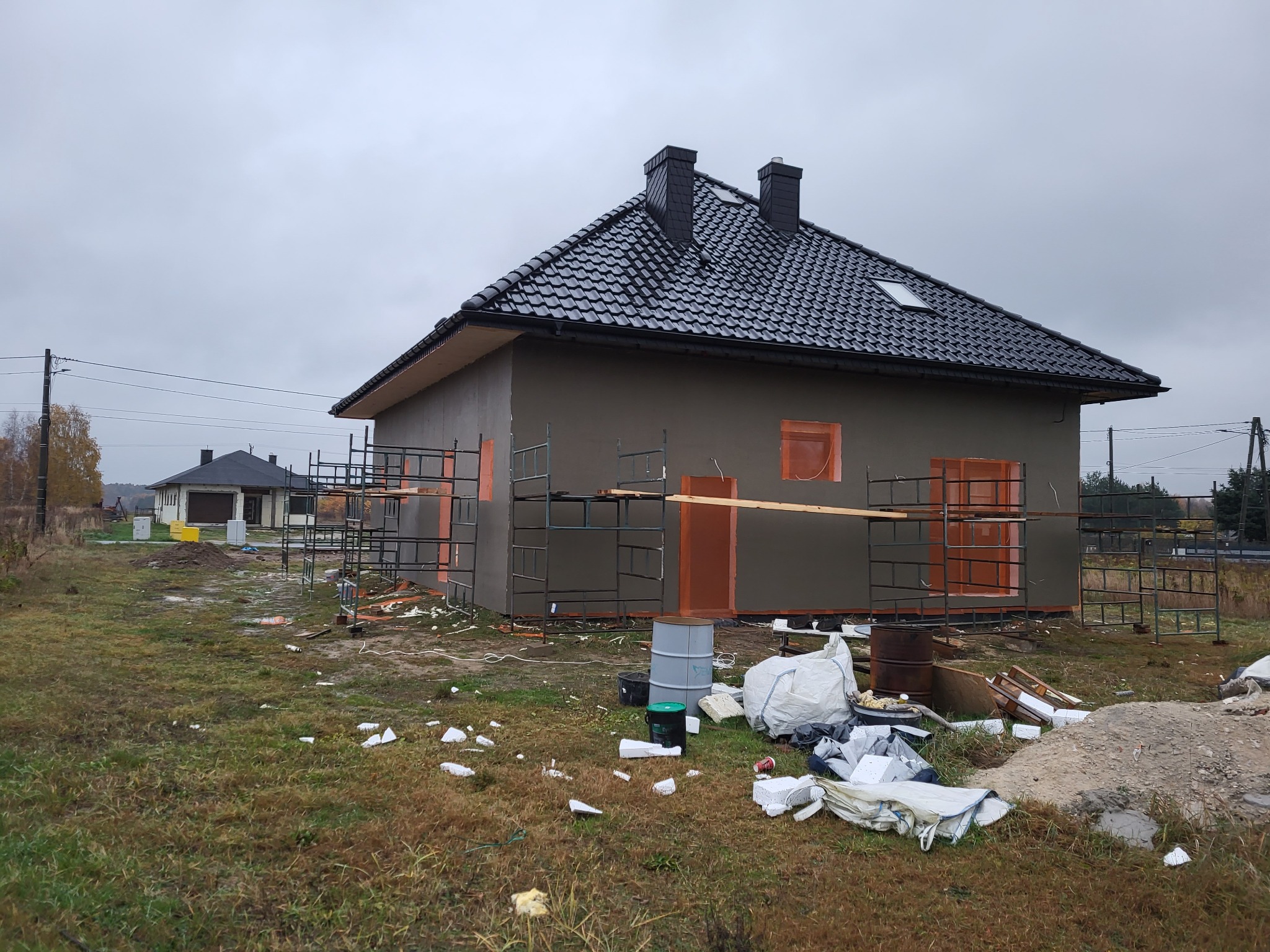 Nowy dom jednorodzinny w trakcie budowy, z czarną dachówką i szarymi ścianami zewnętrznymi, otoczony rusztowaniami i nieporządkiem na trawniku, pochmurne niebo.