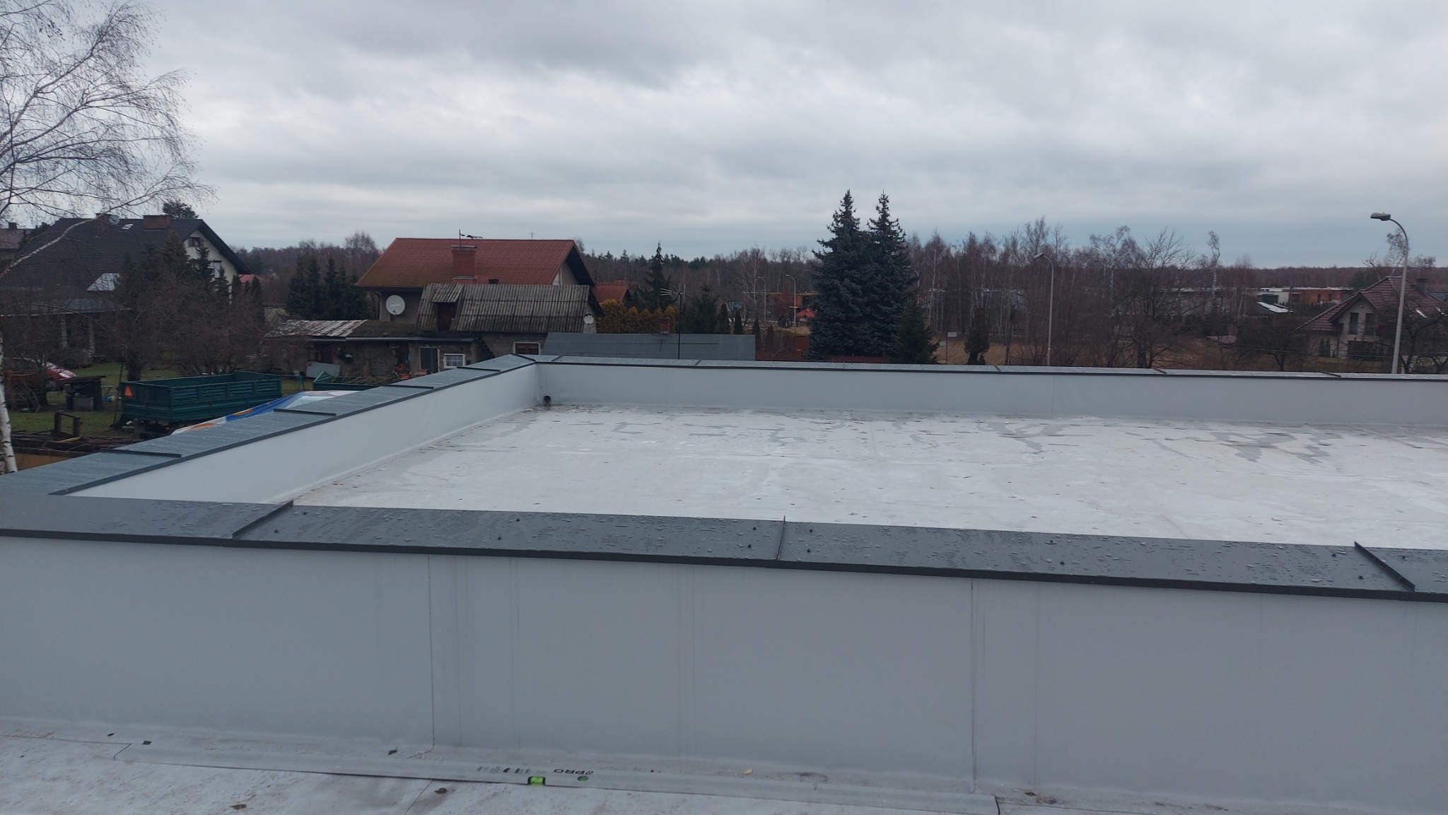Nowoczesny, płaski dach pokryty jasną membraną, z ciemnoszarymi obróbkami blacharskimi na krawędziach. W tle widoczne zabudowania mieszkalne i drzewa pod pochmurnym niebem.
