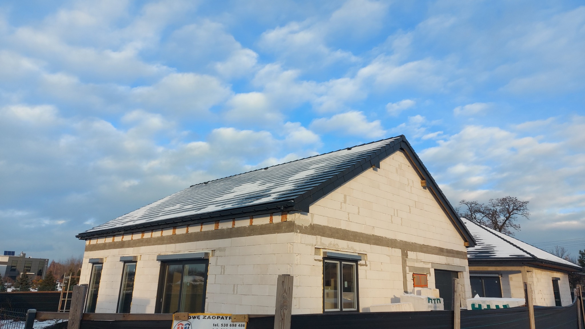 Nowy dom w trakcie budowy z częściowo ośnieżonym, czarnym dachem z dachówki ceramicznej i zamontowanymi rynnami, widoczne niezakończone elewacje z białego bloczka i rolety zewnętrzne.