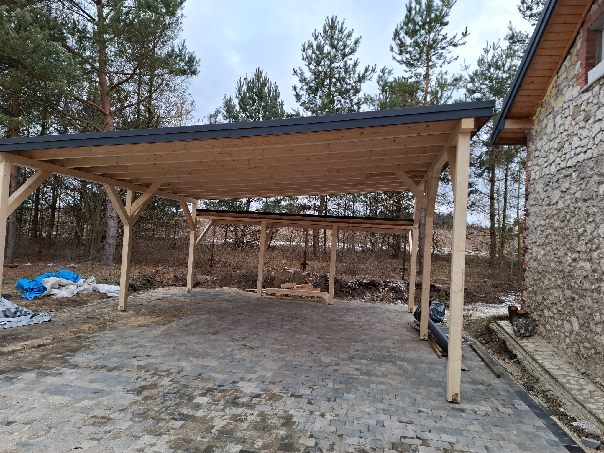 Drewniana konstrukcja z płaskim, ciemnym dachem, prawdopodobnie w trakcie budowy, z widocznymi elementami konstrukcyjnymi i materiałami budowlanymi na ziemi, przylegająca do kamiennego budynku...