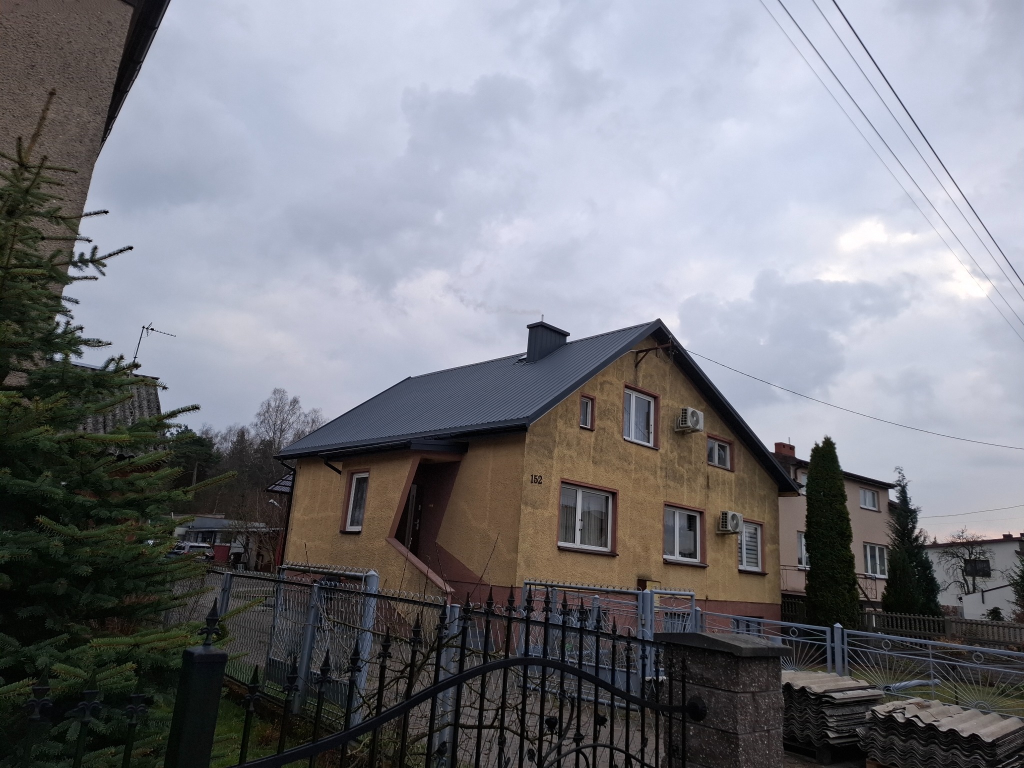 Dom dwukondygnacyjny z nowym, ciemnoszarym dachem blaszanym, widoczny zza metalowego ogrodzenia i świerku, pochmurne niebo.