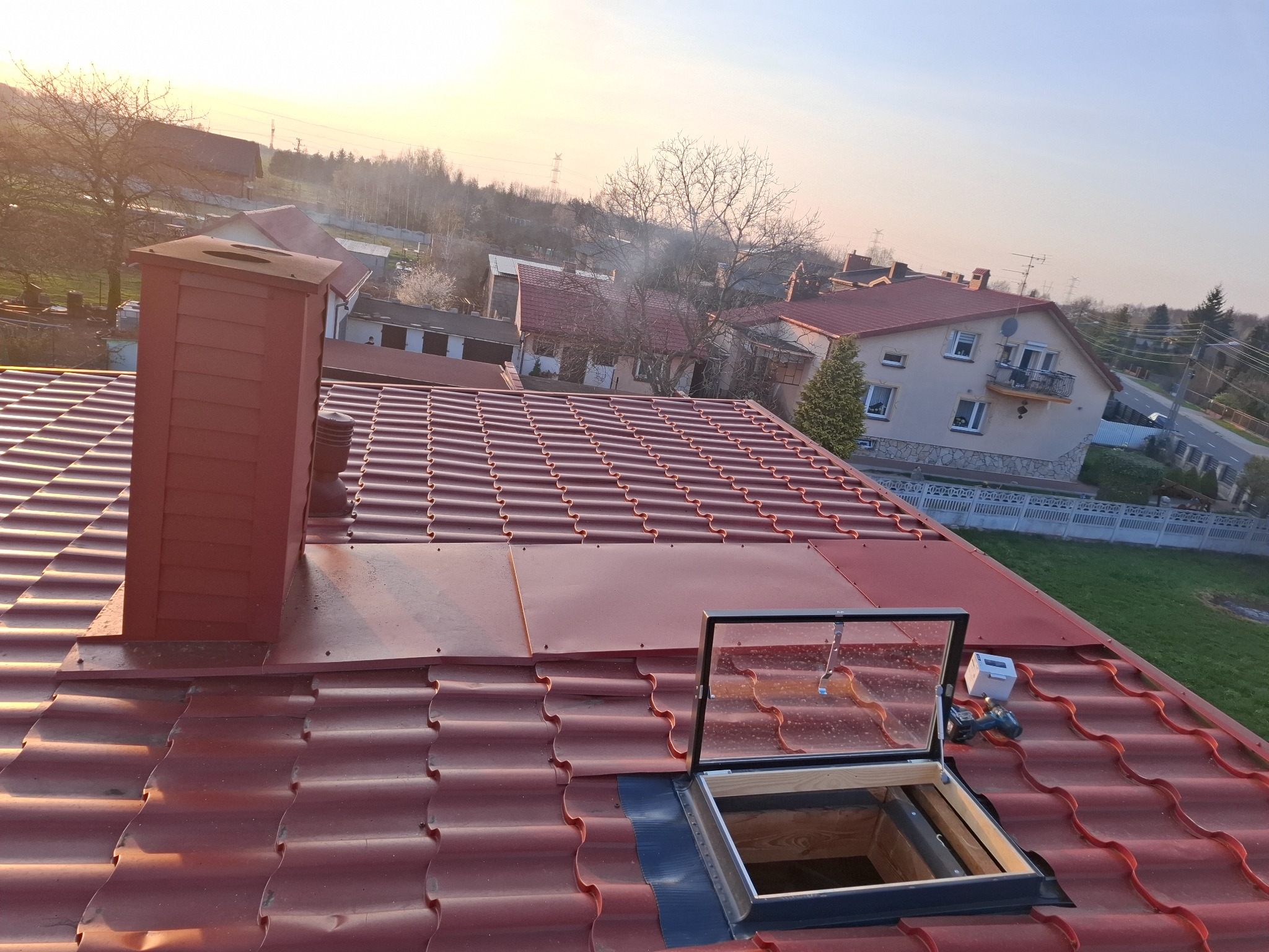 Czerwony dach z dachówki ceramicznej z kominem i otwartym oknem dachowym w trakcie montażu, widoczne narzędzia i elementy montażowe, w tle zabudowa jednorodzinna oświetlona zachodzącym słońcem.