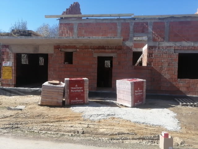 Budowa domu jednorodzinnego z czerwonej cegły Porotherm, widoczne palety z cegłami na placu budowy, w tle niezadaszona konstrukcja dachu.