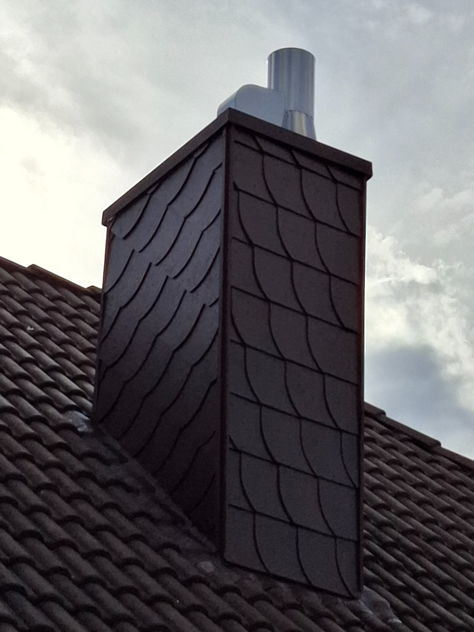 Komin obłożony łupkiem w kształcie rybiej łuski, z metalowym wkładem kominowym, na tle pochmurnego nieba.
