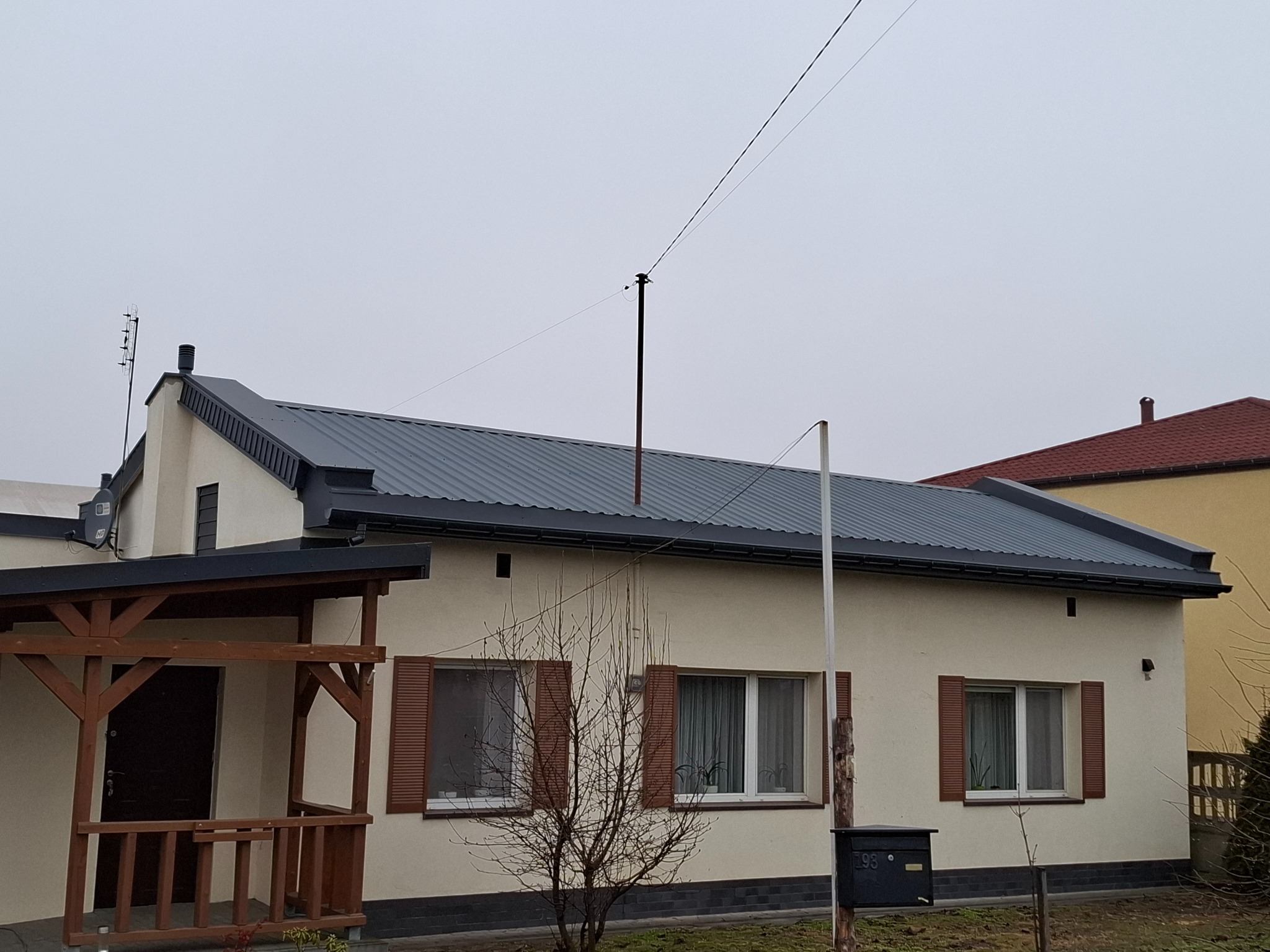 Dom jednorodzinny z nowym, szarym dachem z blachy trapezowej, brązowe okiennice, drewniana weranda i antena na dachu w pochmurny dzień.