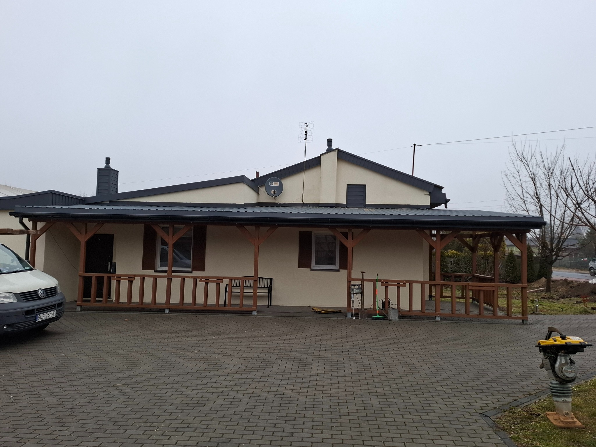 Parterowy dom z ciemnym dachem i drewnianą werandą, widoczny samochód dostawczy i zagęszczarka gruntu.