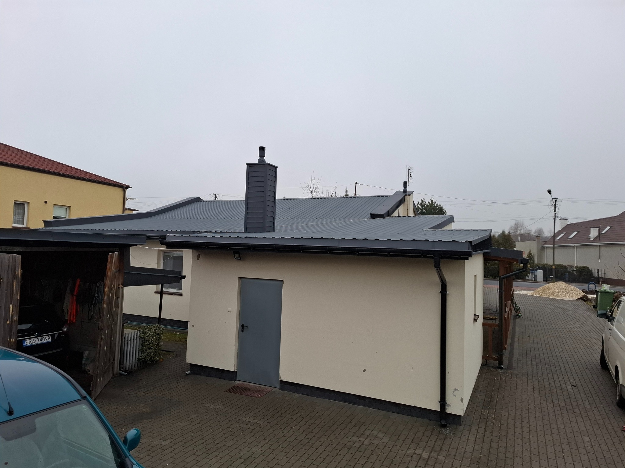 Nowoczesny budynek gospodarczy z ciemnoszarą blachą na dachu i kominem, widoczny z zewnątrz na tle pochmurnego nieba.