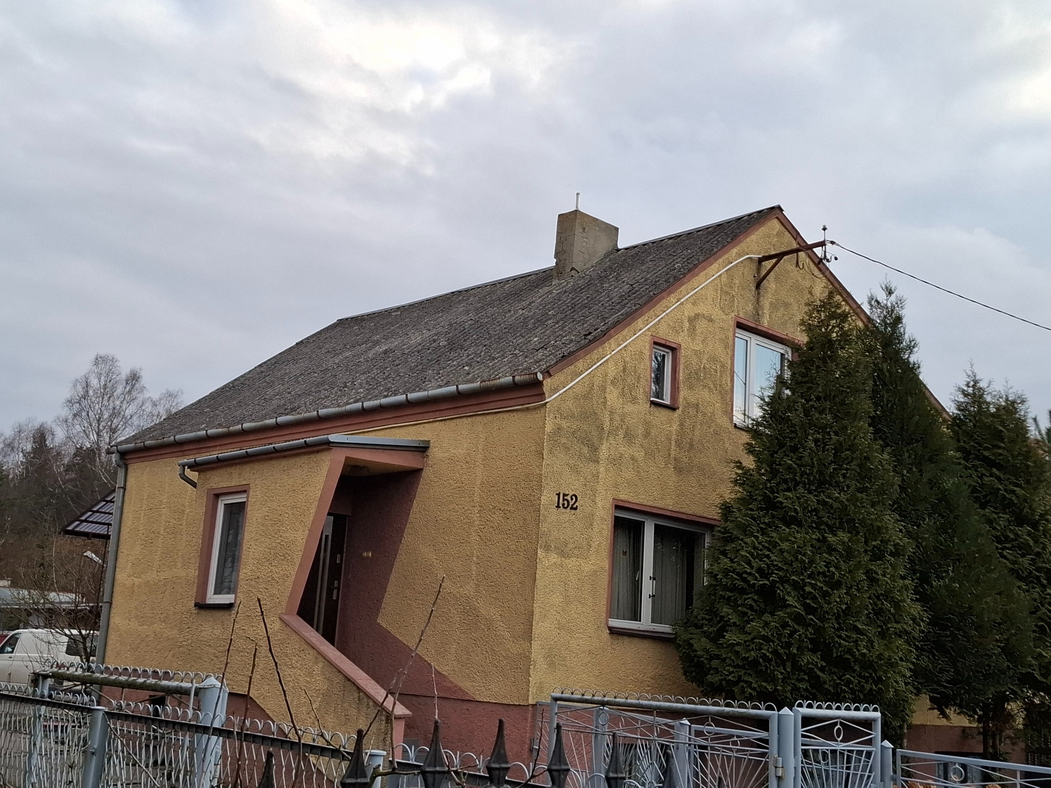 Dom jednorodzinny z szarym dachem krytym eternitem, otynkowany na żółto z brązowymi akcentami, widoczny numer domu '152', otoczony metalowym ogrodzeniem i drzewami iglastymi, pochmurne niebo w tle.