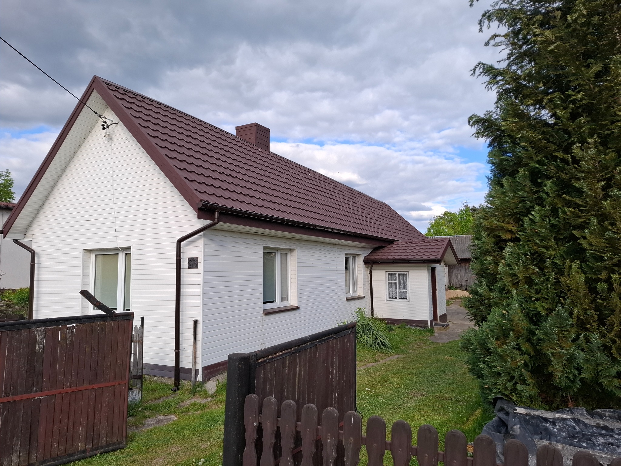Dom jednorodzinny z brązowym dachem z blachodachówki, białą elewacją i brązowymi rynnami, widoczny zza drewnianego płotu. Pochmurne niebo w tle.