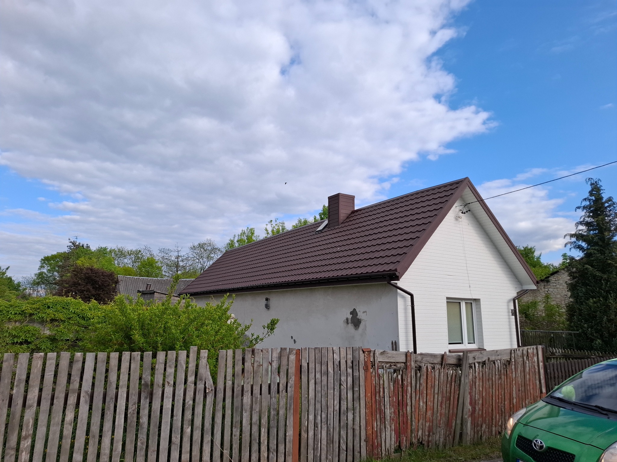 Dom z brązowym dachem krytym dachówką, białą elewacją i drewnianym płotem w tle błękitnego nieba z chmurami. Widok zza ogrodzenia, fragment zielonego auta w kadrze.