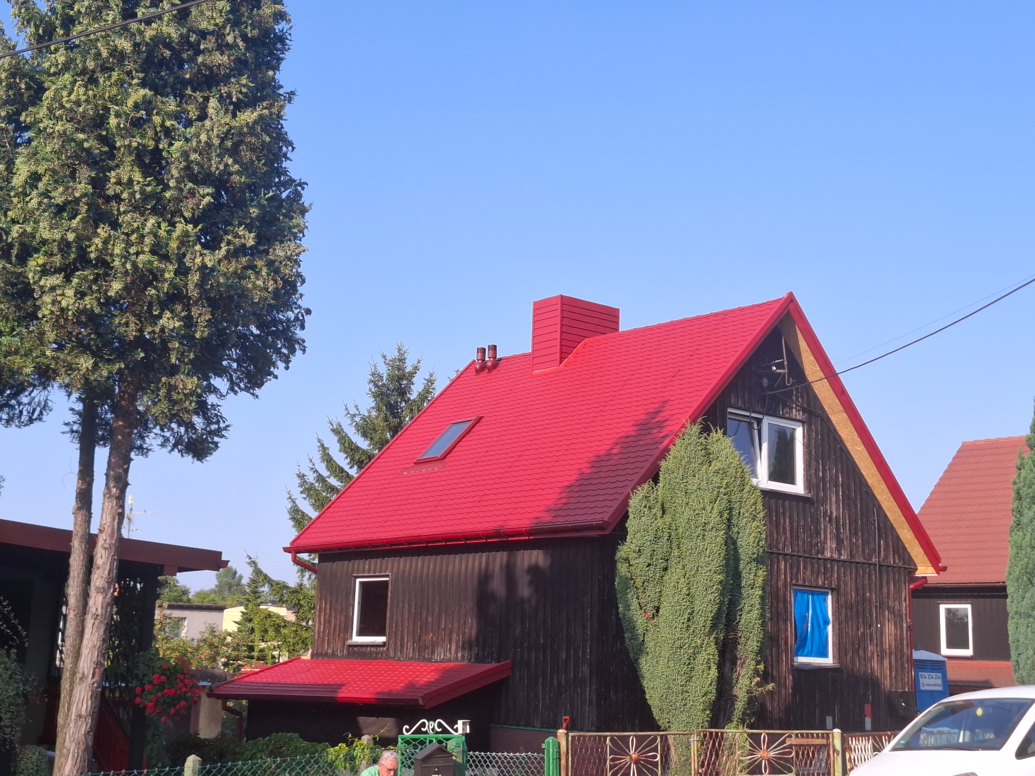 Ciemny dom z nowym, intensywnie czerwonym dachem i oknem dachowym. Widoczne obróbki blacharskie i rynny. Tło stanowi błękitne niebo i zieleń drzew.