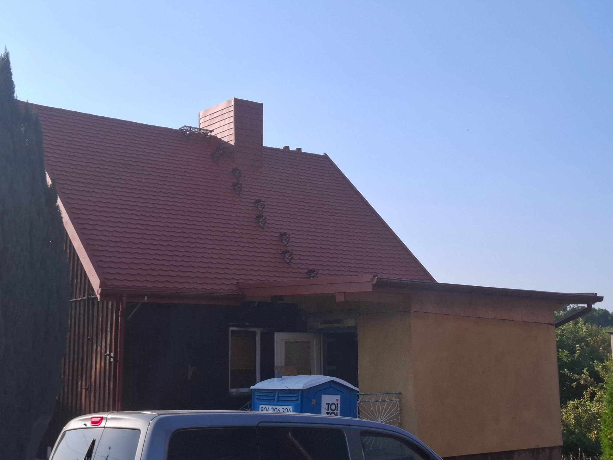 Czerwony dach z blachodachówki na domu w Rędzinach, widoczny z dołu. Fragment samochodu w kadrze. Dach z kominem i zabezpieczeniami przeciwśniegowymi.