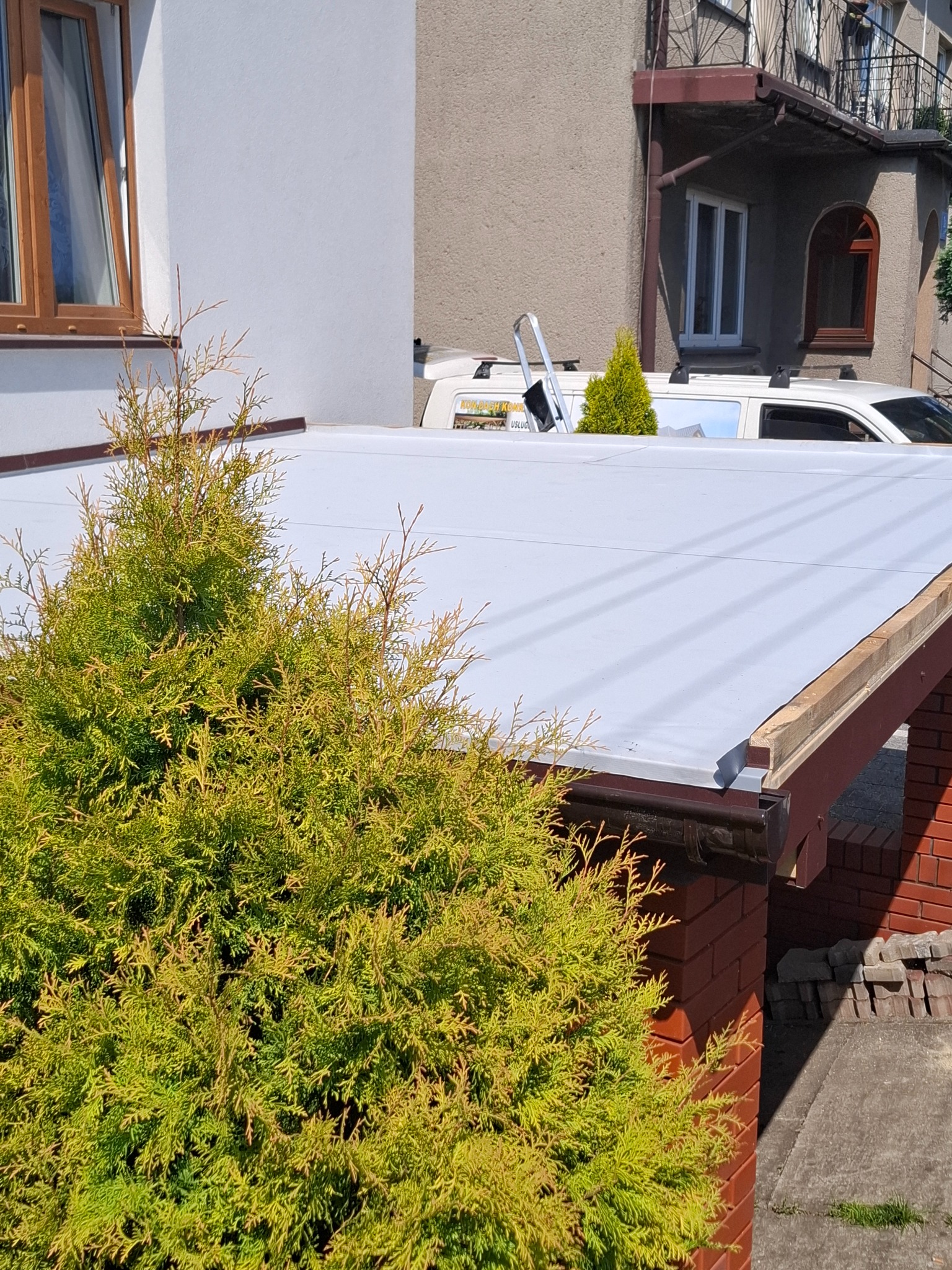 Nowe, jasnoszare pokrycie dachu płaskiego z brązowymi rynnami, widoczne spod zielonych krzewów, z samochodem firmy dekarskiej w tle.