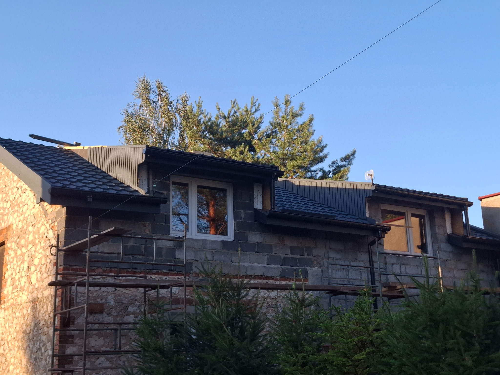 Dom w trakcie prac dekarskich: częściowo nowy, czarny dach z blachy, fragment ściany z rusztowaniem. Widoczne drzewa i błękitne niebo.