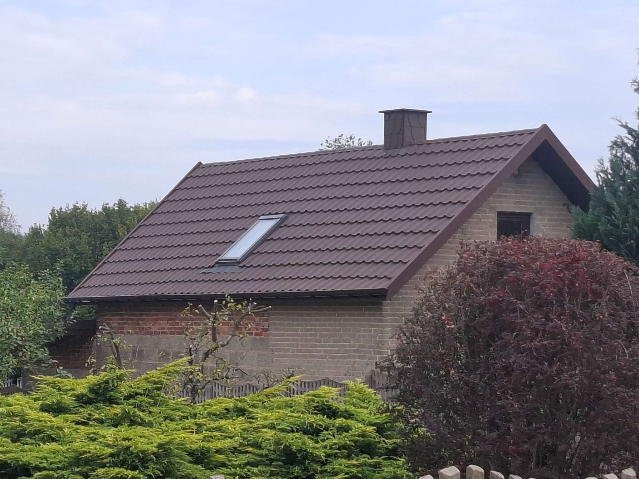 Dom z brązowym dachem z dachówki trapezowej i oknem dachowym, komin z obróbką blacharską. Budynek otoczony zielenią w słoneczny dzień.