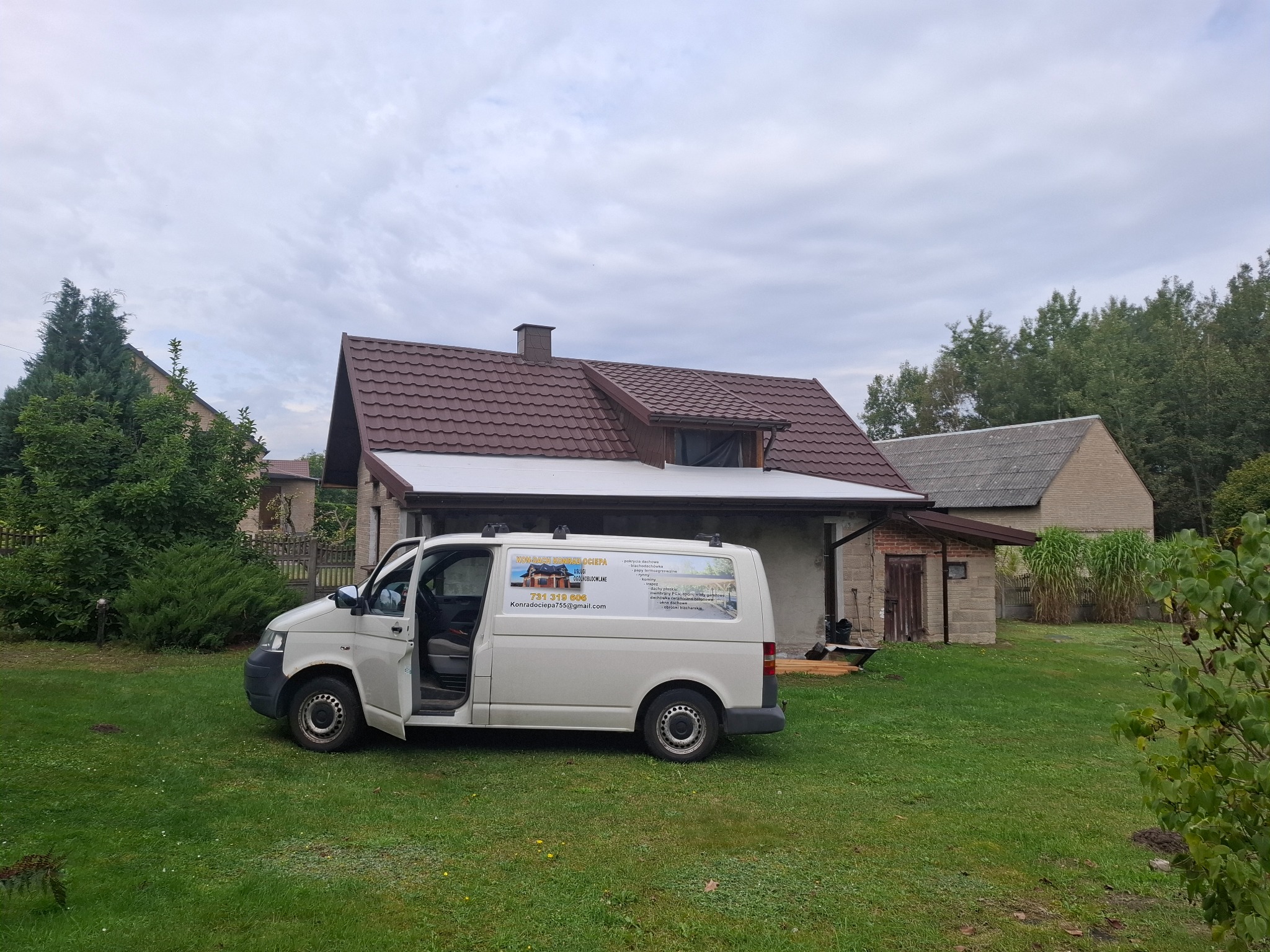 Dom z brązowym dachem, obok biały bus z reklamą usług dekarskich: pokrycia dachowe, obróbki blacharskie, kominy. Zieleń trawnika i drzew kontrastuje z pochmurnym niebem.