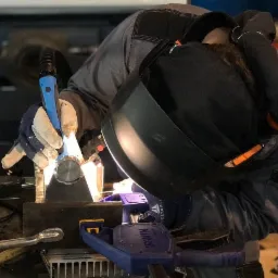 Pracownik w kombinezonie ochronnym i masce spawalniczej, podczas spawania metalowego elementu zamocowanego w zaciskach, z użyciem uchwytu spawalniczego z niebieskim elementem.