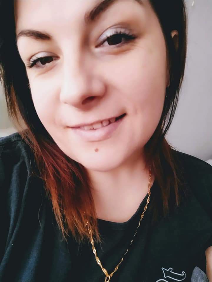 Uśmiechnięta kobieta z pieprzykiem na twarzy i złotym łańcuszkiem, prezentująca się w czarnej koszulce.