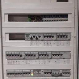 Rozdzielnica elektryczna w białej obudowie z widocznymi bezpiecznikami, listwami zaciskowymi i przewodami, z zieloną diodą sygnalizacyjną na jednym z wyłączników.