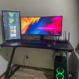 Stanowisko komputerowe z dwoma monitorami, klawiaturą podświetlaną RGB, myszką, routerem Wi-Fi i obudową komputera z podświetleniem LED, umieszczone na czarnym biurku o metalowych nogach na tle...