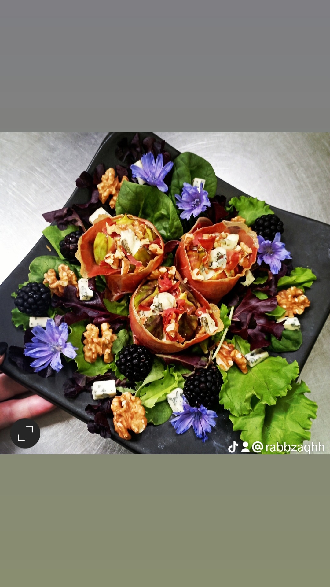 Elegancko podana sałatka z szynką parmeńską, serem pleśniowym, orzechami włoskimi, jeżynami i jadalnymi kwiatami na czarnym, kwadratowym talerzu.