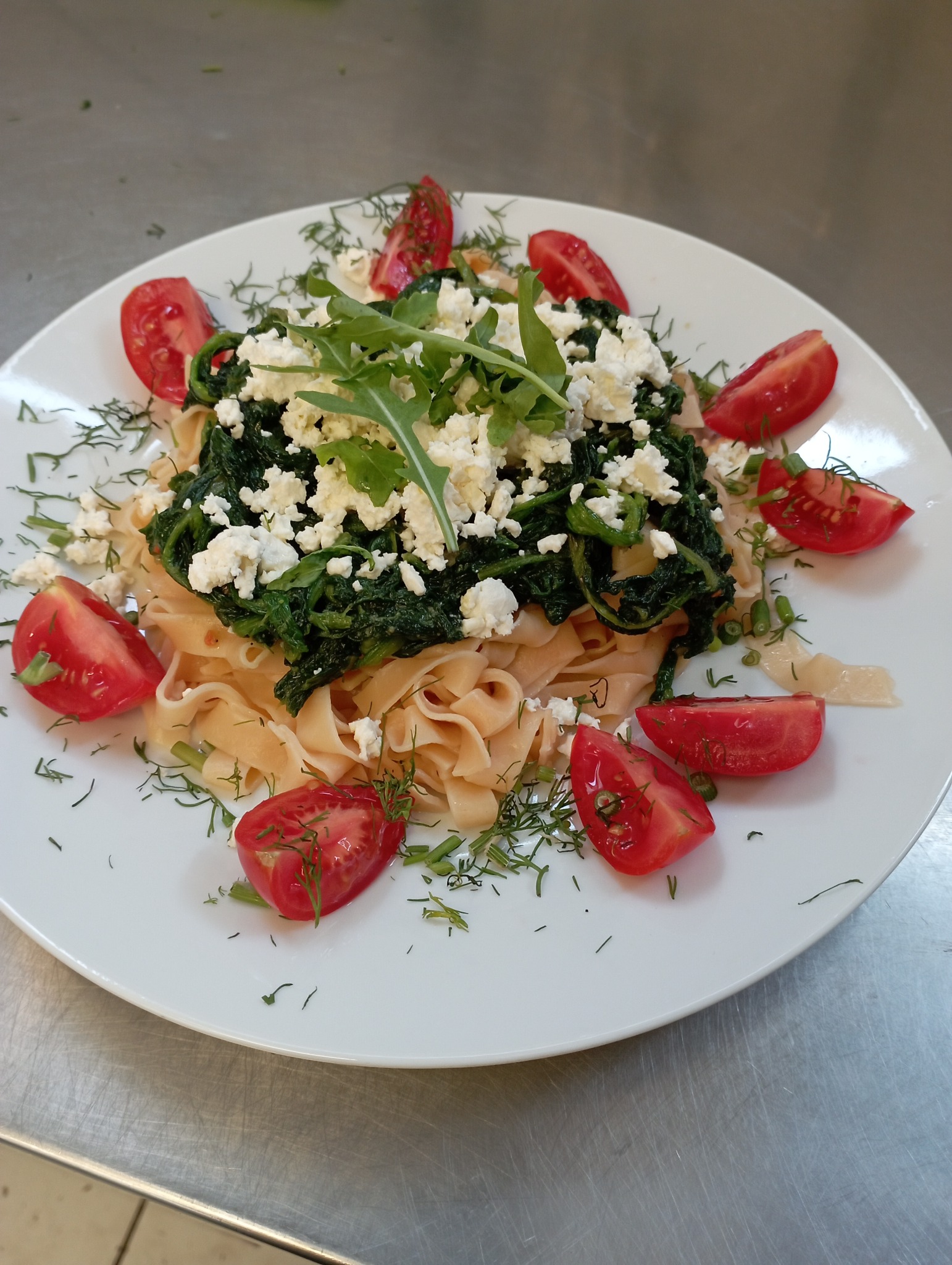 Tagliatelle ze szpinakiem, serem feta, pomidorami i świeżym koperkiem na białym talerzu, ujęcie z góry na tle metalowego blatu.