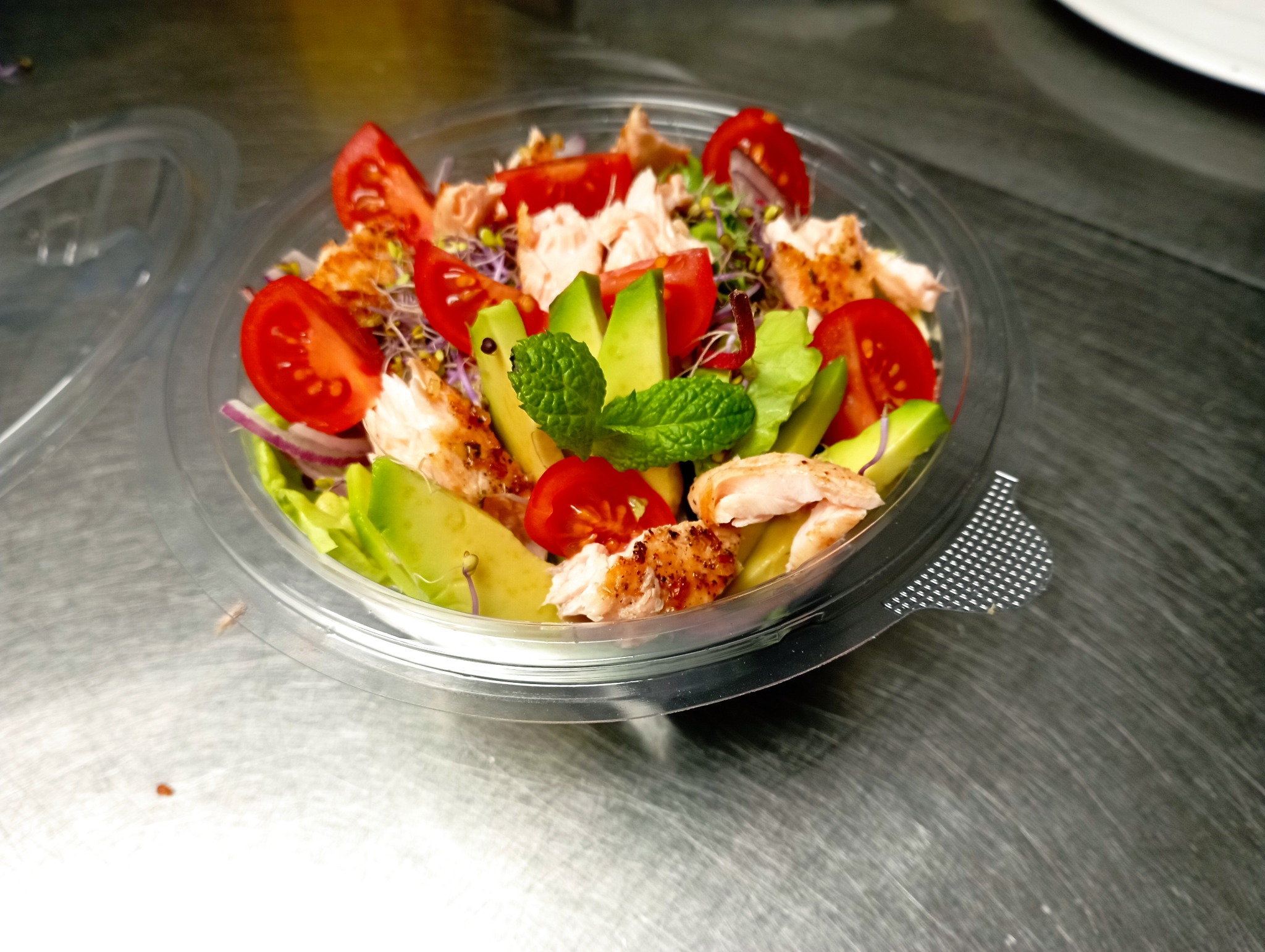 Sałatka z grillowanym kurczakiem, awokado, pomidorkami koktajlowymi, czerwoną cebulą i kiełkami w przezroczystym plastikowym pojemniku, udekorowana listkiem mięty, na metalowym blacie.