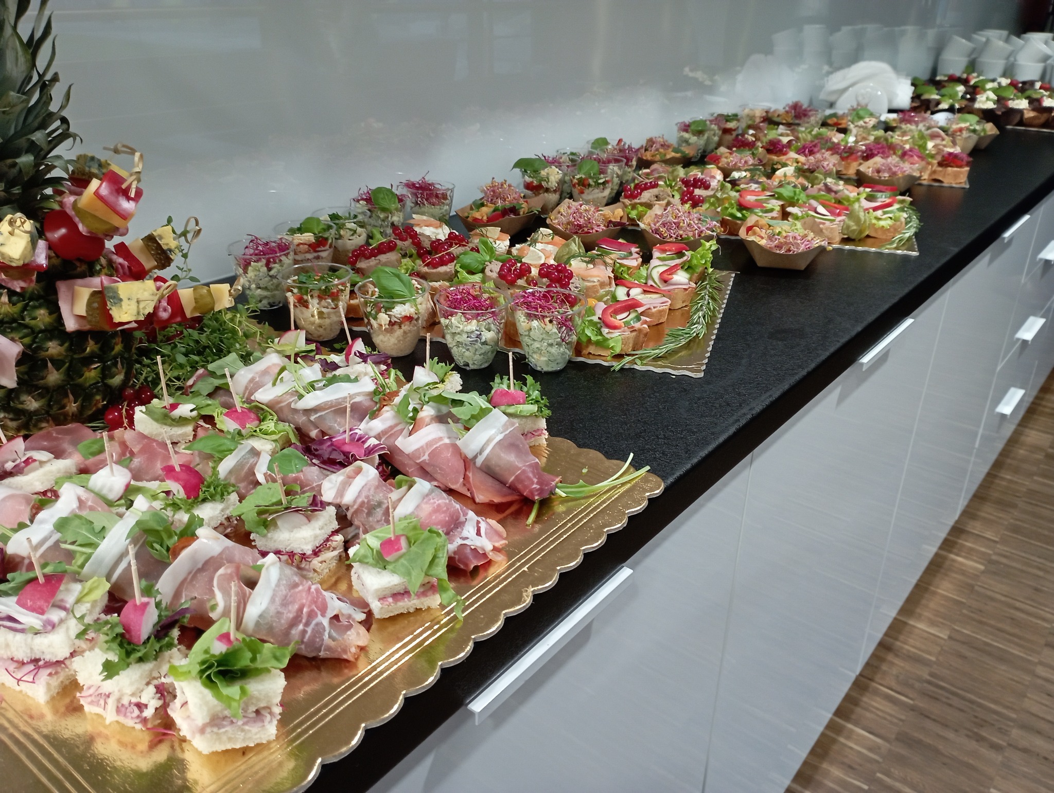 Długi stół z czarnym blatem, na którym znajdują się różnorodne, apetyczne przekąski: kanapeczki z szynką parmeńską i rzodkiewką, sałatki w małych szklankach i mini-tarty z owocami, a na końcu...