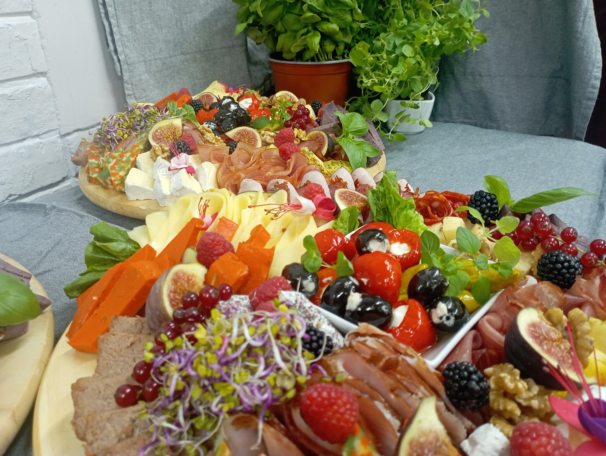 Dwa okrągłe, drewniane półmiski z różnorodnymi przekąskami: sery, wędliny, owoce (figi, maliny, jeżyny, czerwona porzeczka), papryczki faszerowane, kiełki, orzechy włoskie, zioła, na szarym tle...