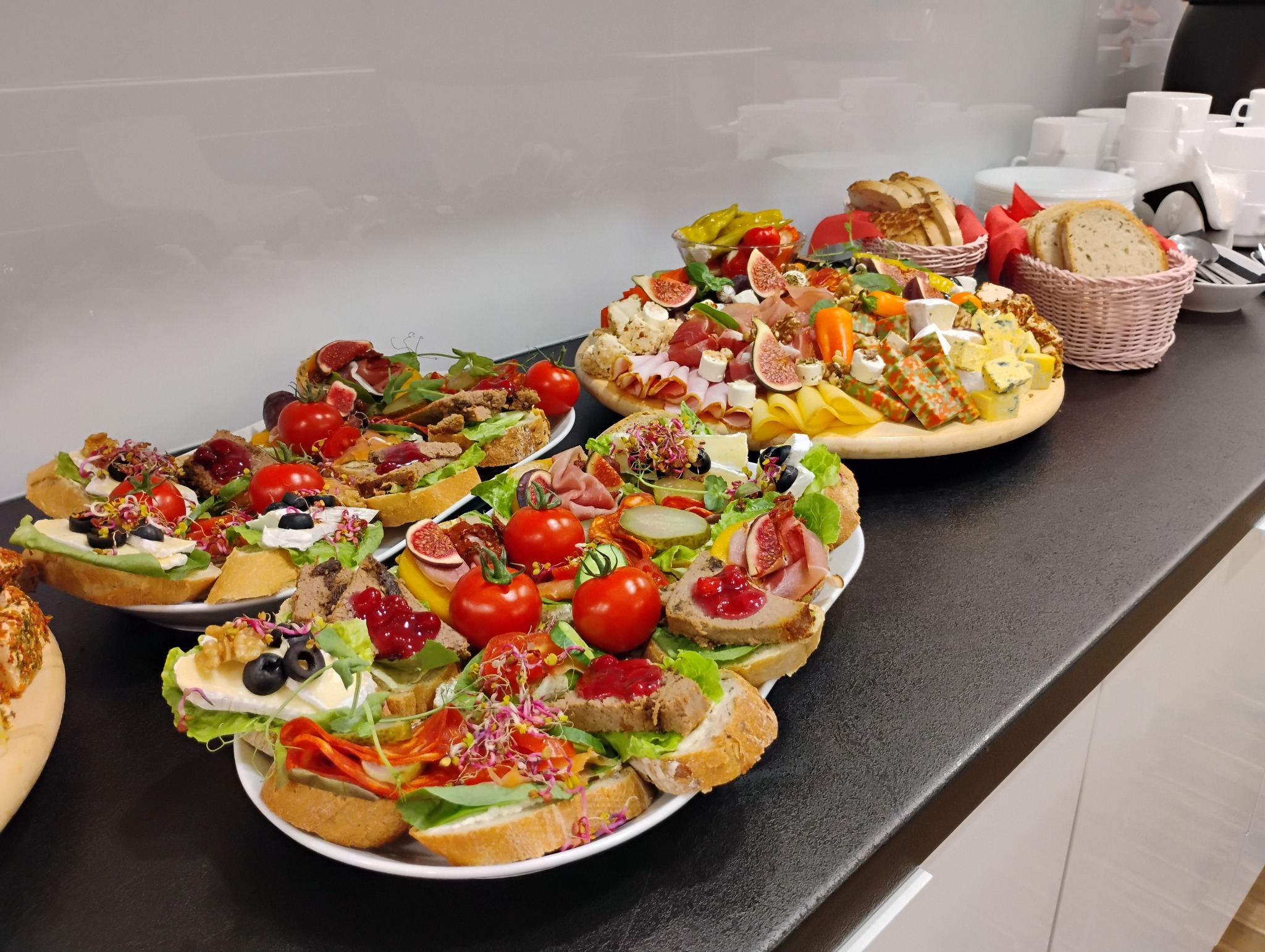 Ułożone na stole talerze i deski z kanapkami, przekąskami i pieczywem, z figami, pomidorami, wędlinami, serami i warzywami, przygotowane do serwowania.