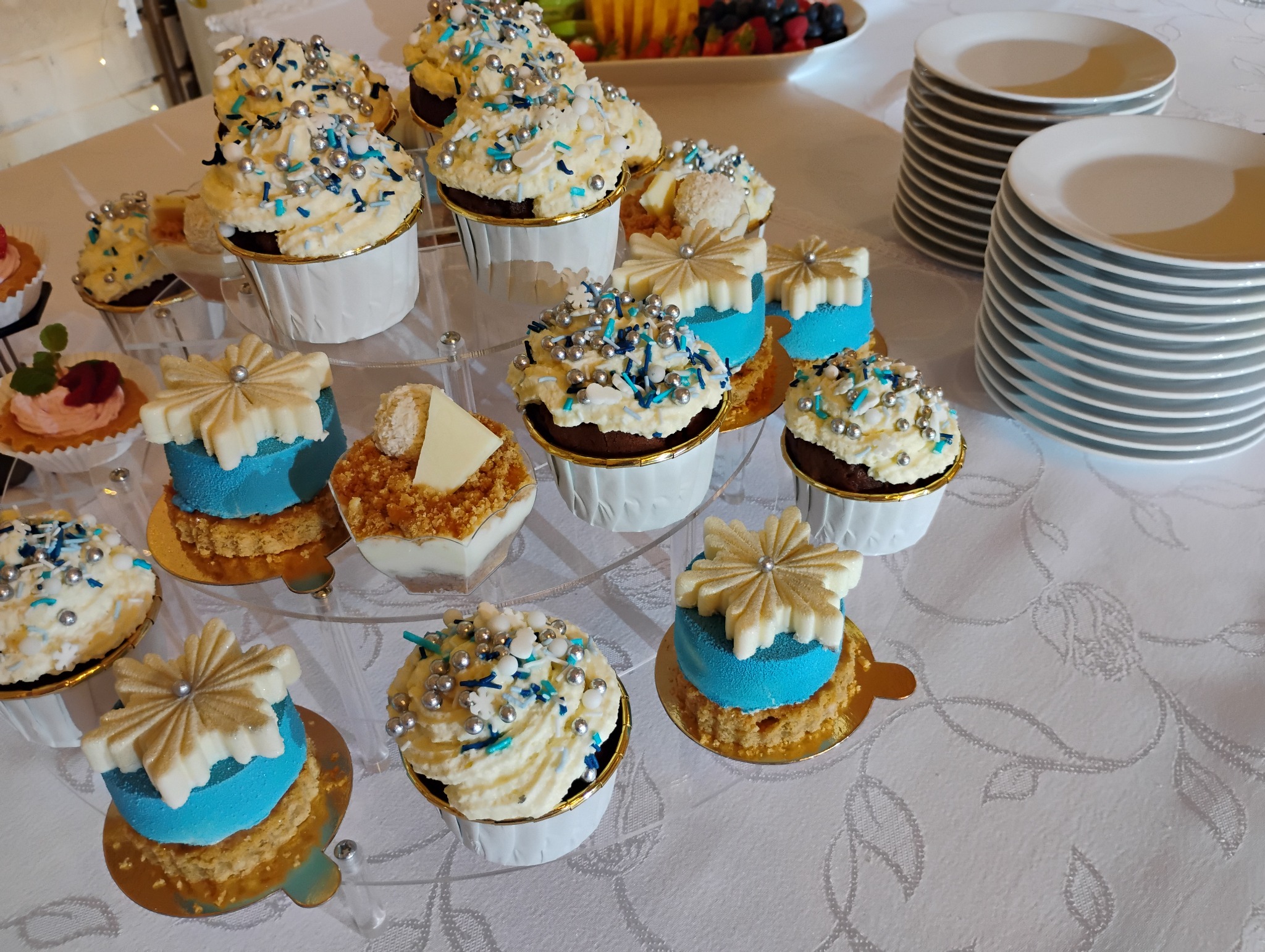 Dekoracyjne babeczki i mini torty na paterach w Przeczycach, z kremem, posypką i dekoracjami w kształcie śnieżynek, obok stosy białych talerzy.