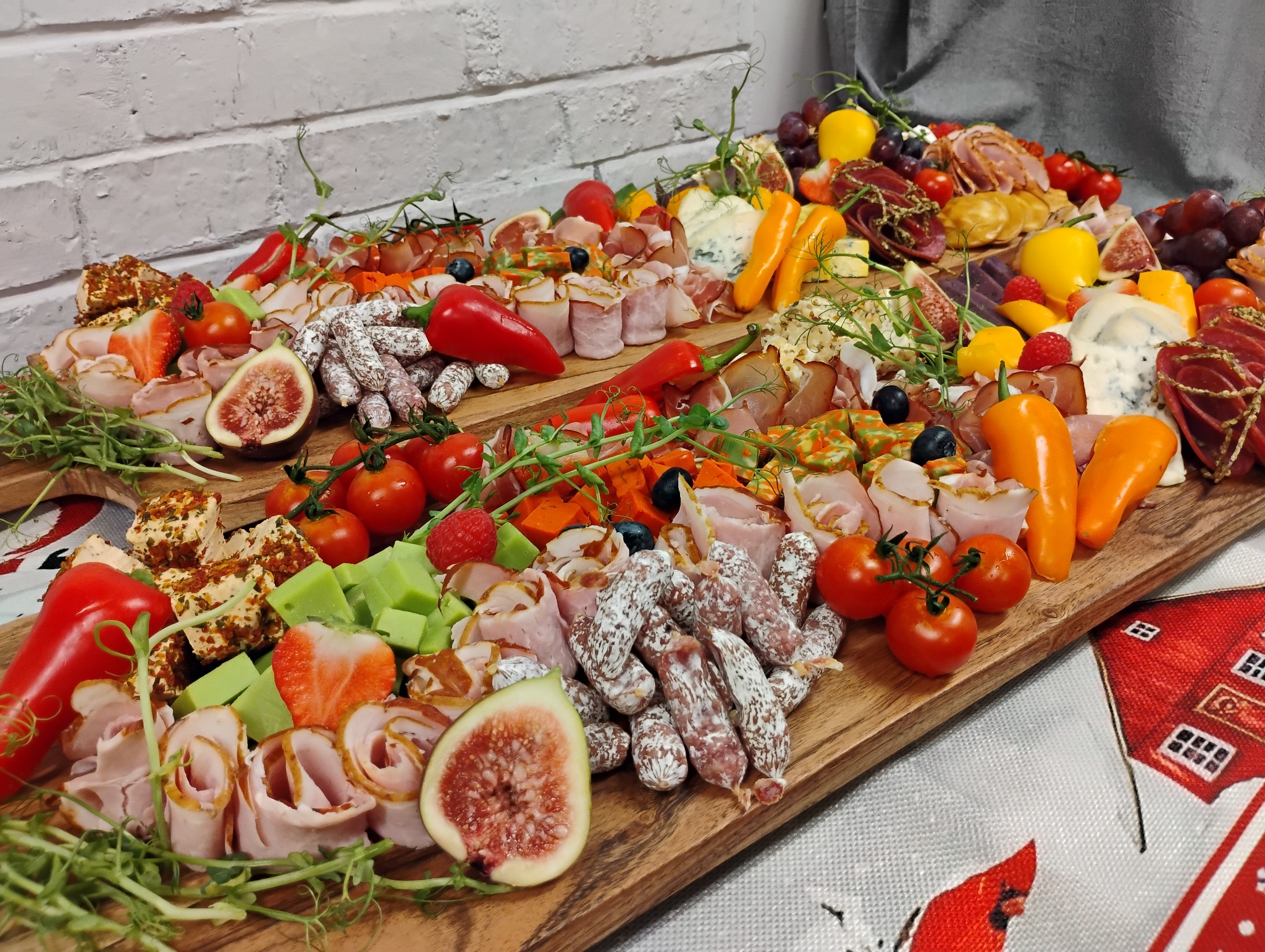 Bogato zaaranżowana deska serów i wędlin z figami, mini paprykami, winogronami i pomidorkami koktajlowymi, ułożona na drewnianym blacie z białym murem w tle.